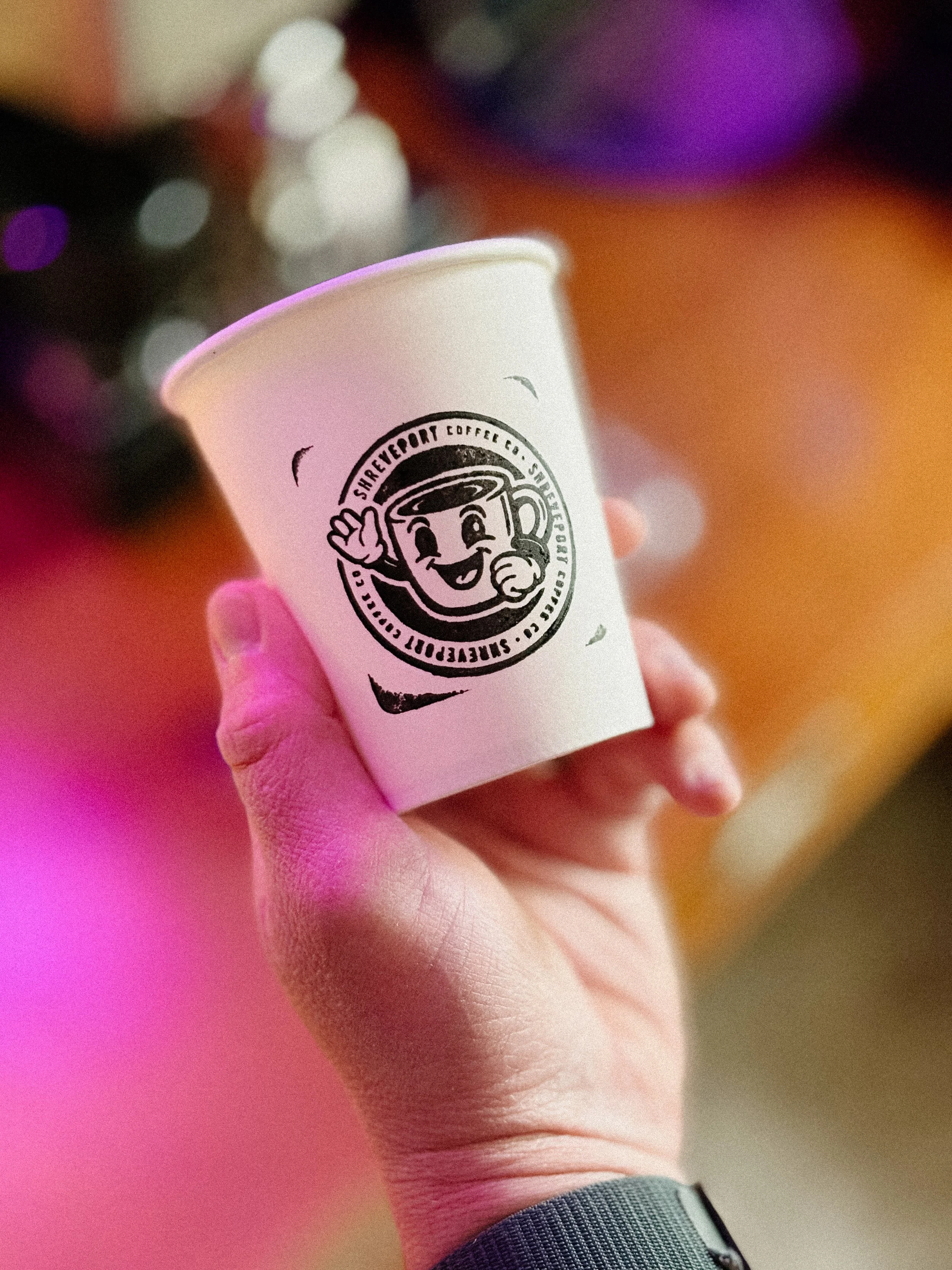 Close-up of a hand holding a white paper cup with a black logo, with a blurred colorful background.