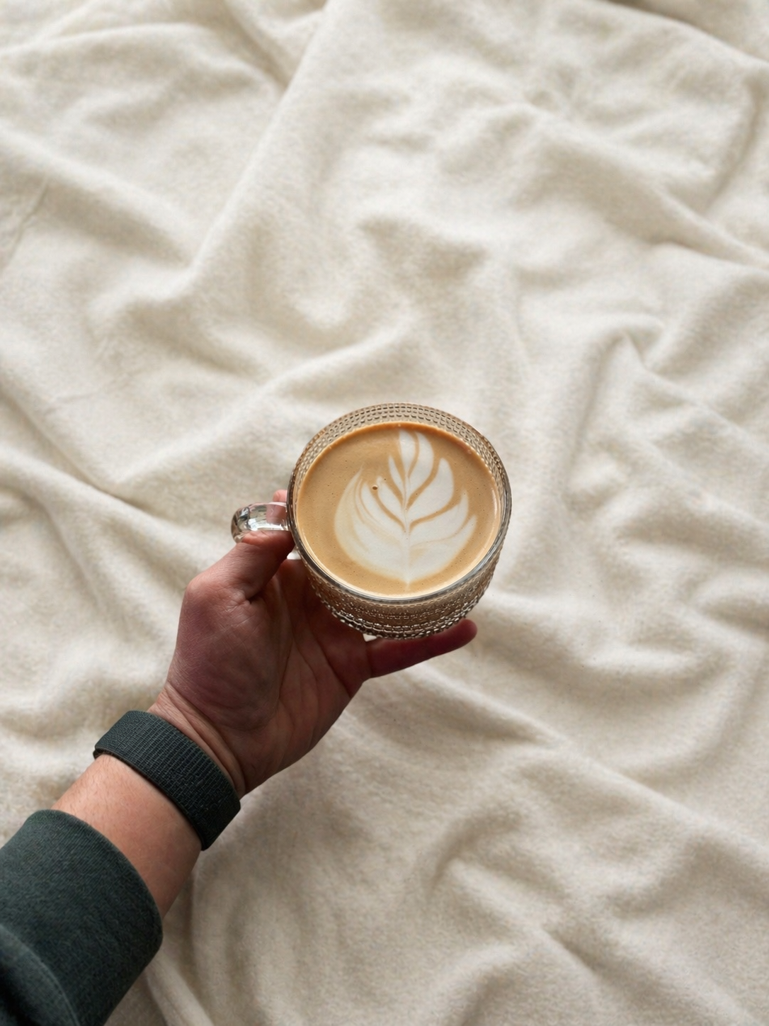 A hand holding a glass mug of coffee with latte art, resting on a cream-colored, wrinkled fabric background.