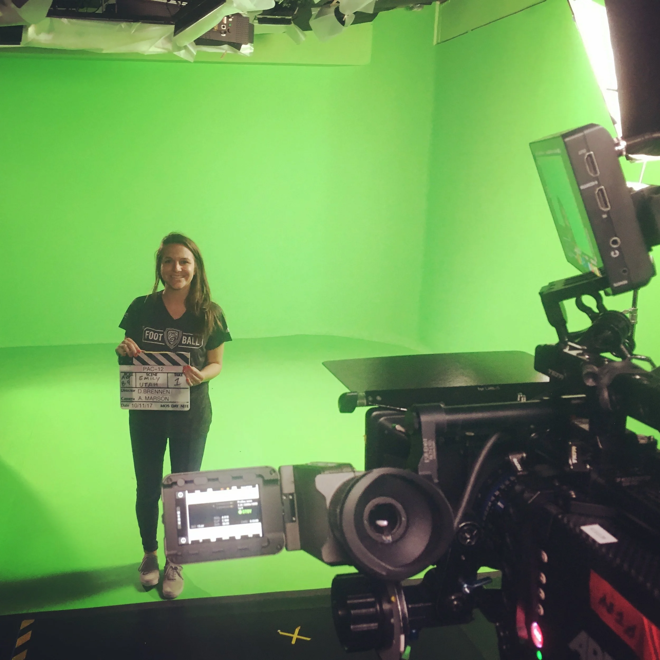 A woman standing in front of a green screen, holding a film slate. Behind her is filming equipment.