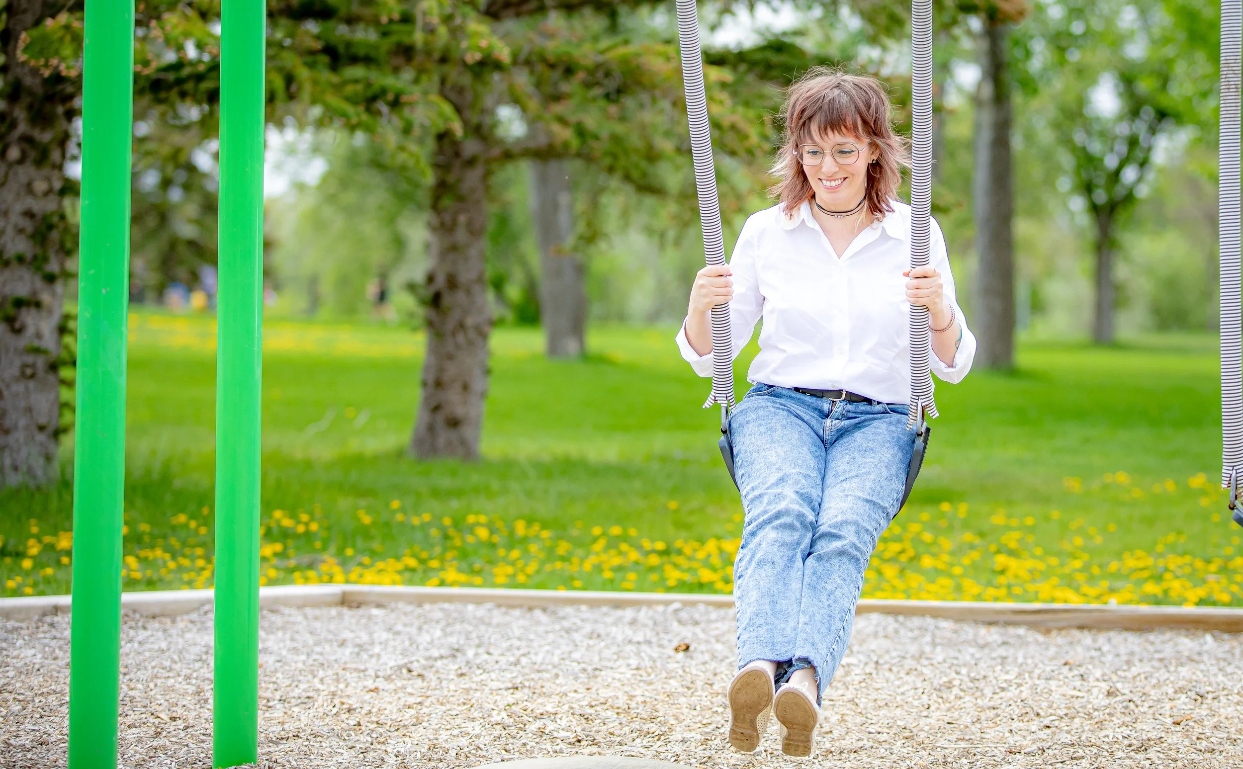 A young woman with glasses and a choker smiling while sitting on a swing at a park with green trees and blooming yellow flowers in the background.