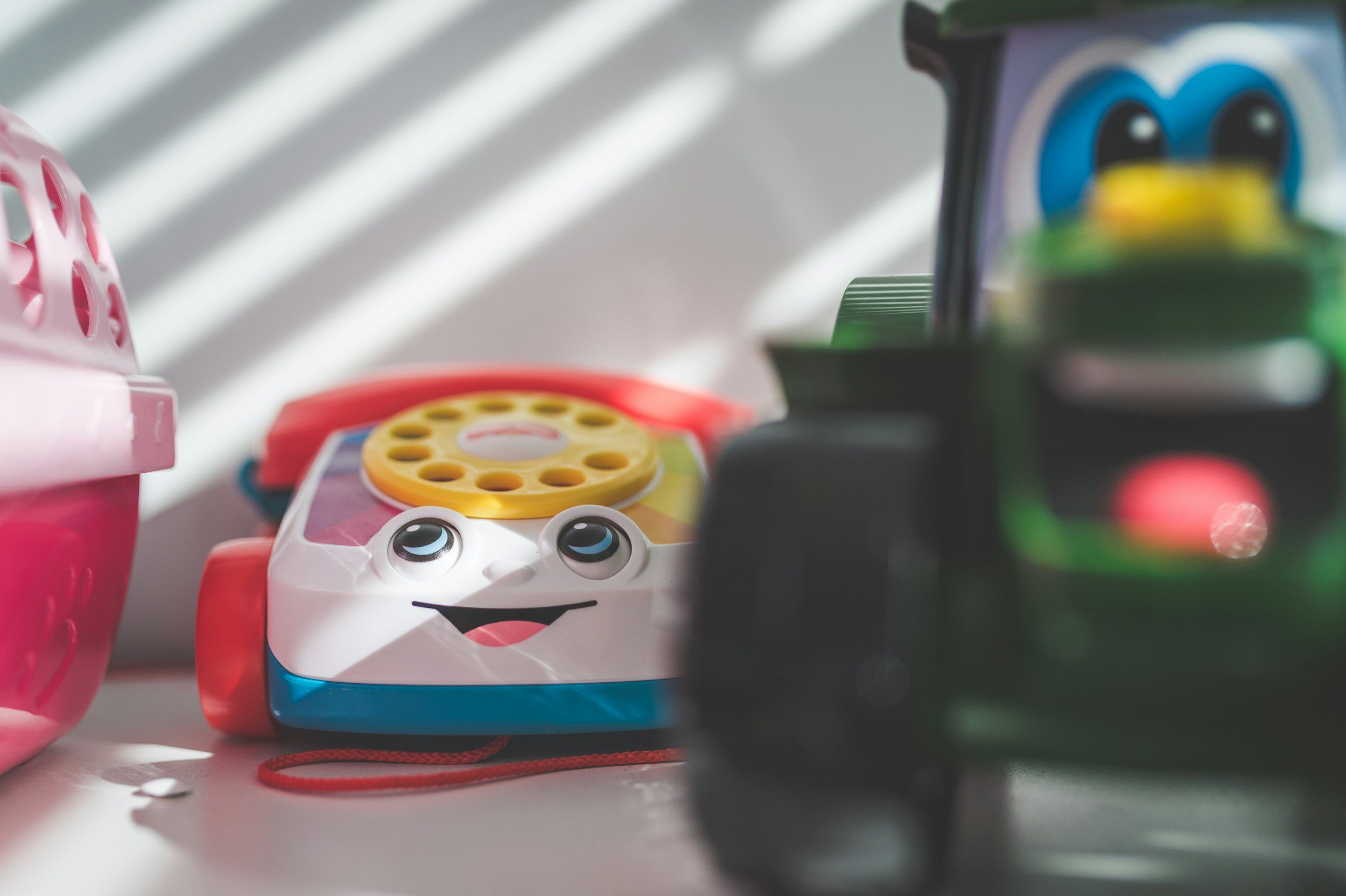 Colorful toy with cartoon face, likely a small device or game, in front of a pink container and other toys or objects on a table, with sunlight creating shadows in the background.