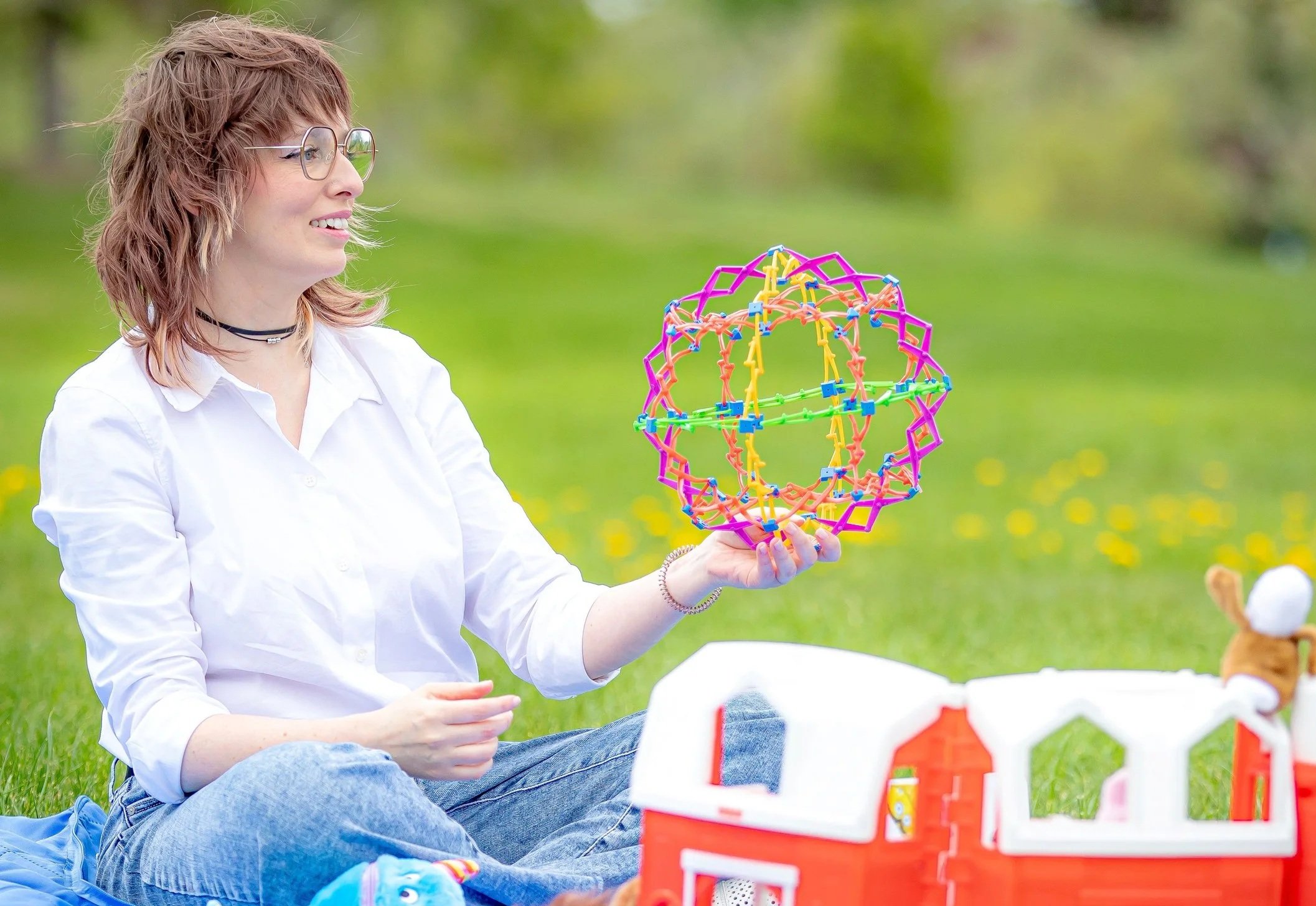 A woman with short hair, glasses, and a white shirt sitting on grass, holding a colorful geometric spherical structure, with a toy farm set in front of her and a blurred green background.