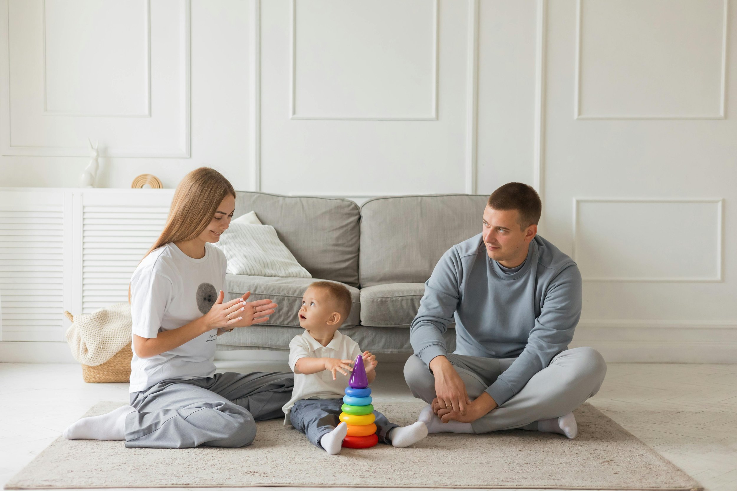 A family of four playing together in a bright, white living room, sitting on a beige rug. The mother, with blonde hair, and the father, with short dark hair, are engaging with a young boy who is stacking colorful rings on a cone toy. The room has a white couch in the background and a minimalist decor style.