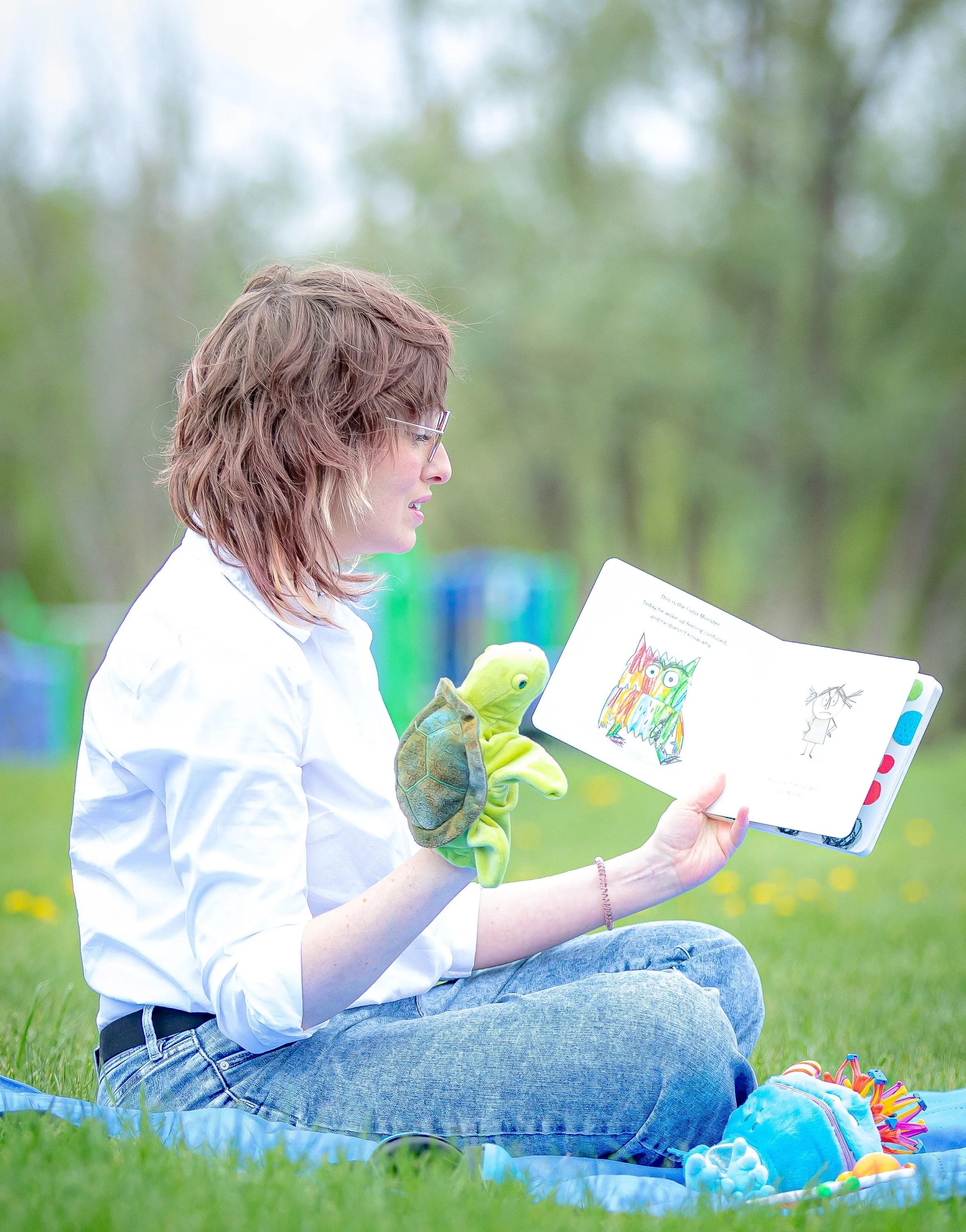 Woman sitting on the grass outdoors reading a children's book with colorful illustrations, a plush turtle toy, and craft supplies nearby.