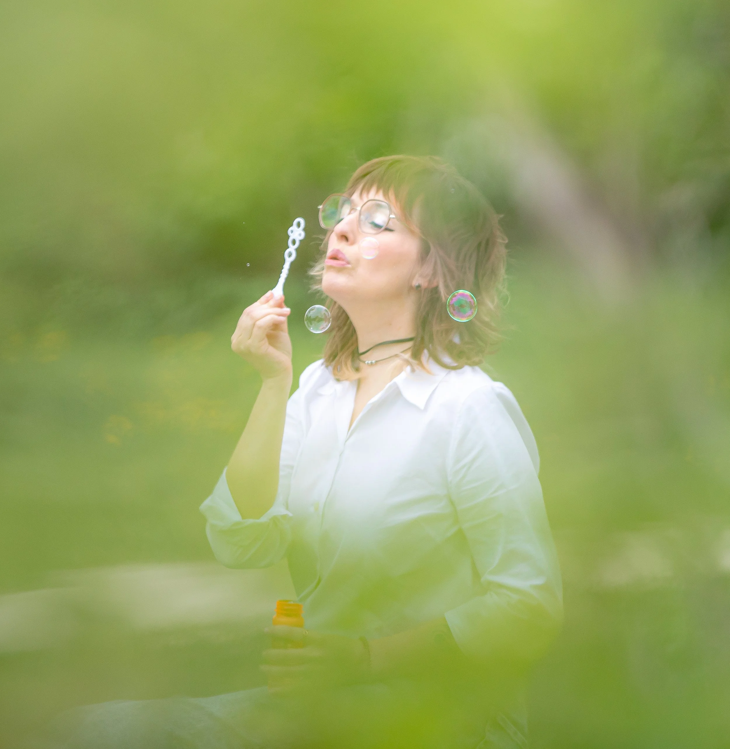 A young woman with glasses and short hair blowing bubbles outdoors with a green blurred background.