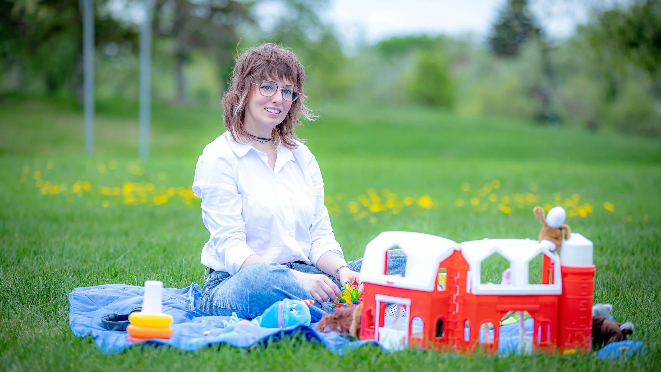 A young woman with wavy brown hair, glasses, and a white shirt sitting on grass in a park with yellow flowers, playing with toys and a dollhouse.