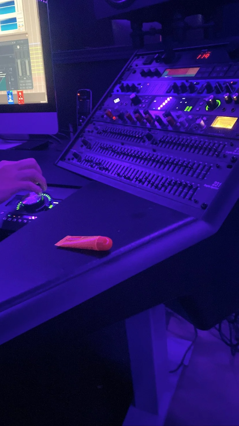 A music production station with a computer monitor, a sound mixing console with various knobs and sliders, a DJ controller with a hand on the jog wheel, and a small tube of lip balm on the desk, illuminated by purple and blue lighting.