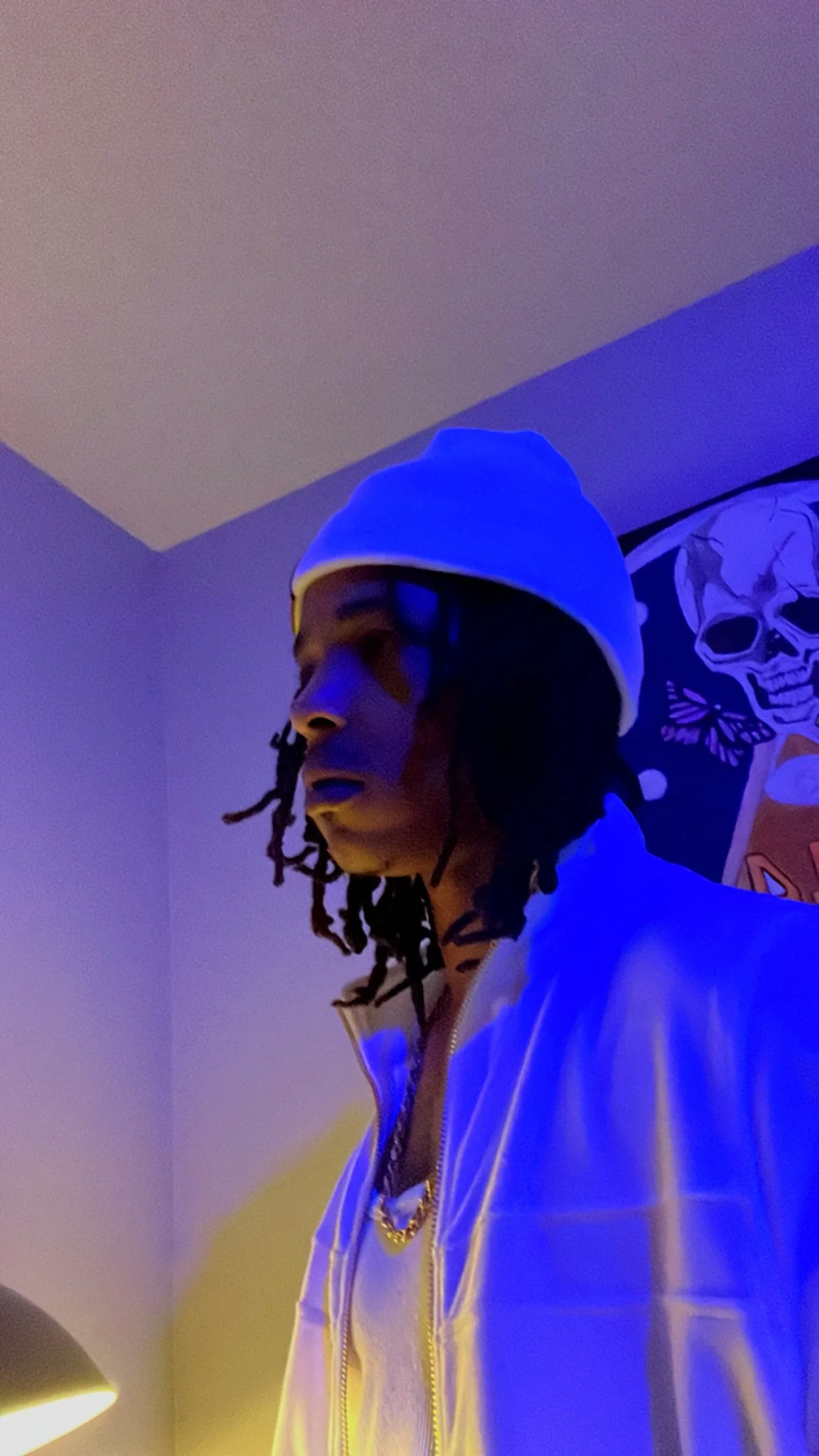 A person with dreadlocks wearing a white hoodie and a beanie, illuminated by blue and yellow lighting, with a skull-themed poster on the wall behind them.