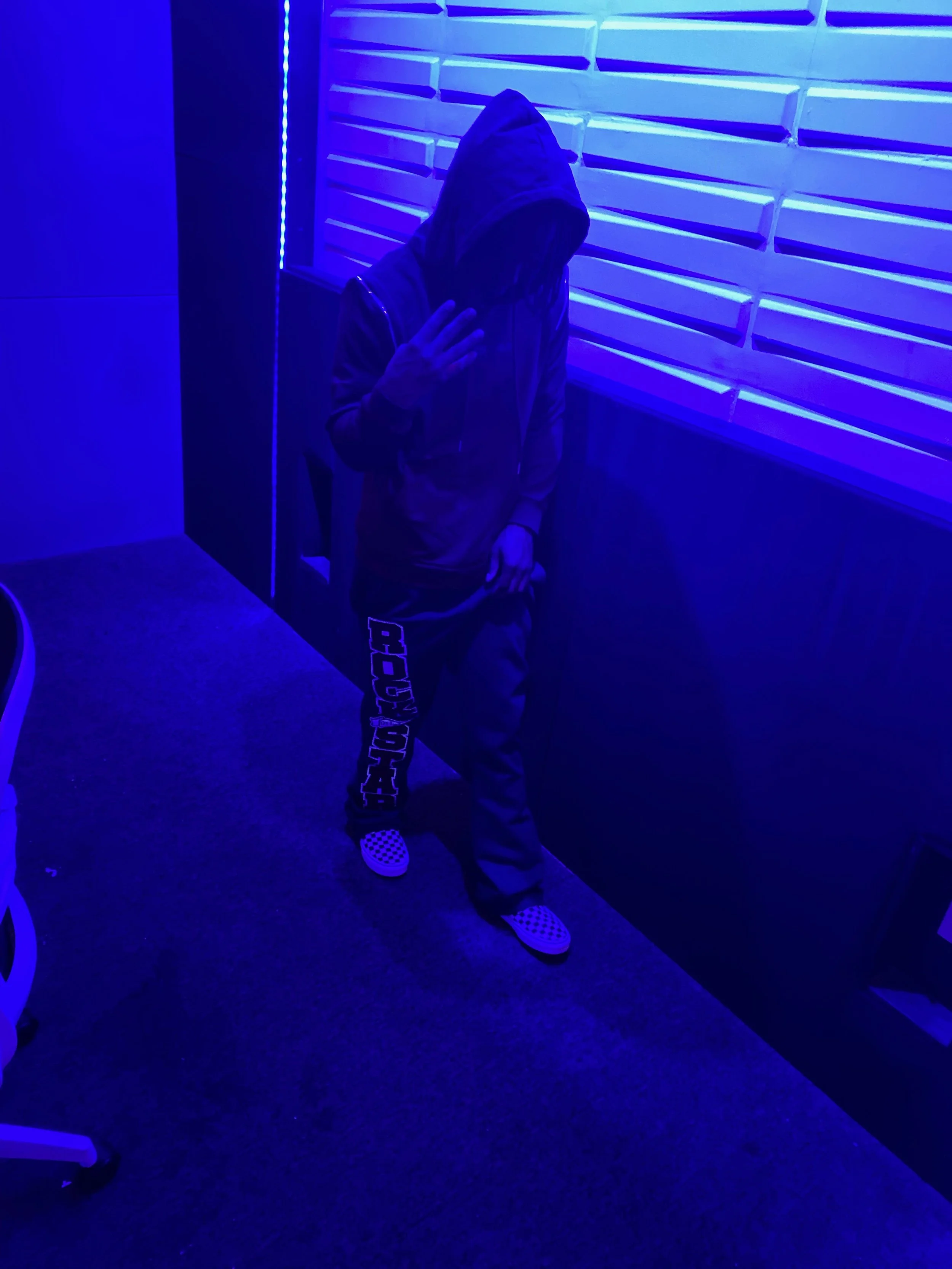Person with dreadlocks wearing a dark hoodie and sweatpants, standing in a room illuminated with blue neon lighting, with horizontal wall panels and an adjacent wall with vertical neon lights.