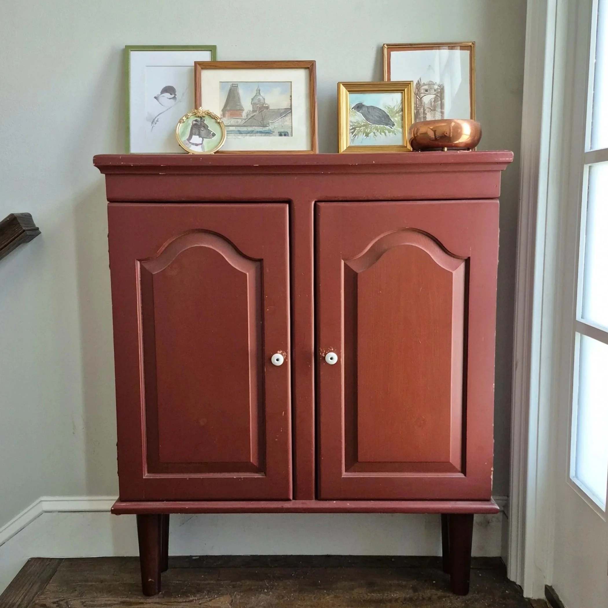 Gold, green, and brown frames of water color paintings sit on a brick red entry console.