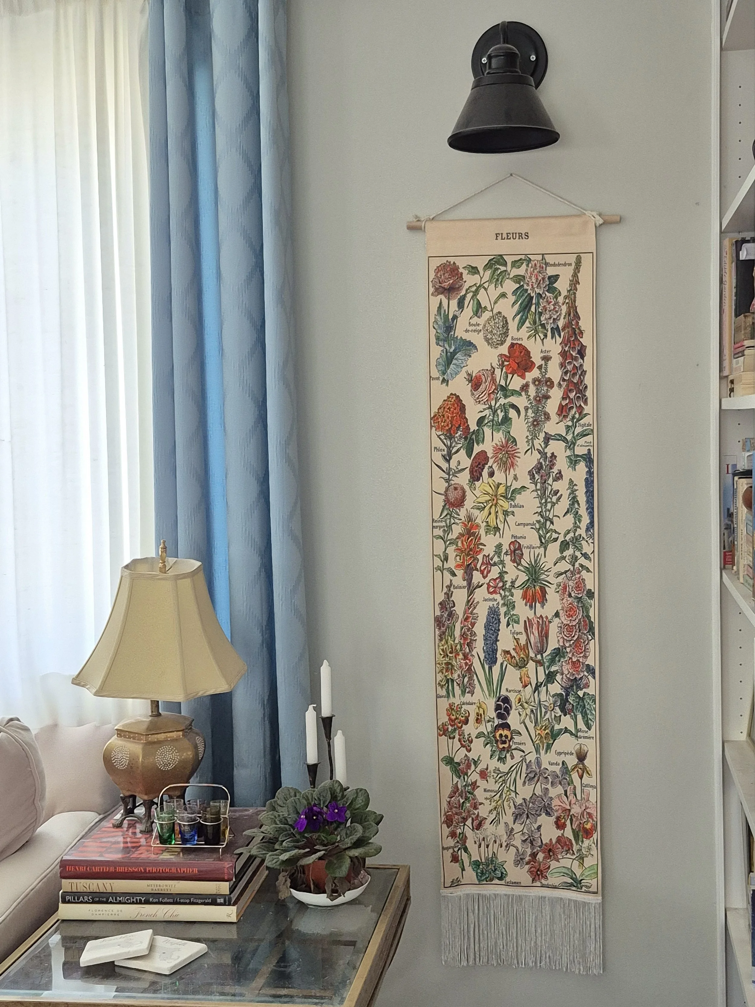 A decorative floral tapestry hangs alongside an end table. The end table has a lamp resting on coffee table books next to candles and an African Violet.