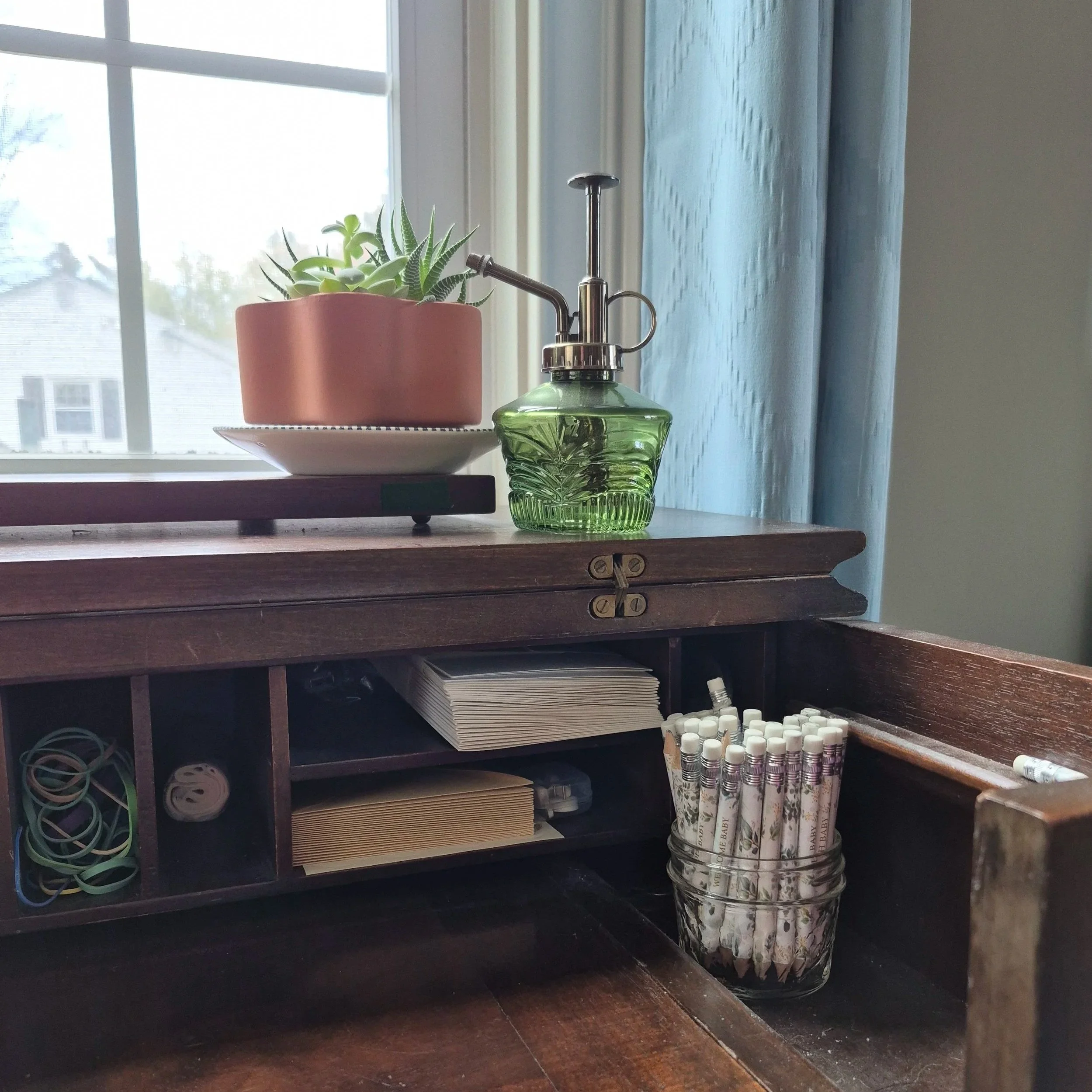 A potted succulent sits next to a water mister atop a spinet desk stocked with stationary and pencils.