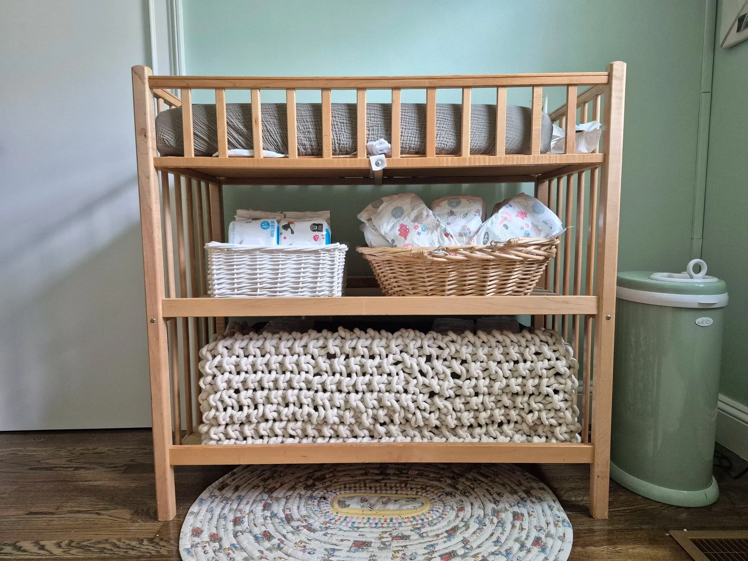 A light wood changing table with green changing pad is fully stocked with a shelf of diapers and wipes, and a shelf with a storage basked beneath.