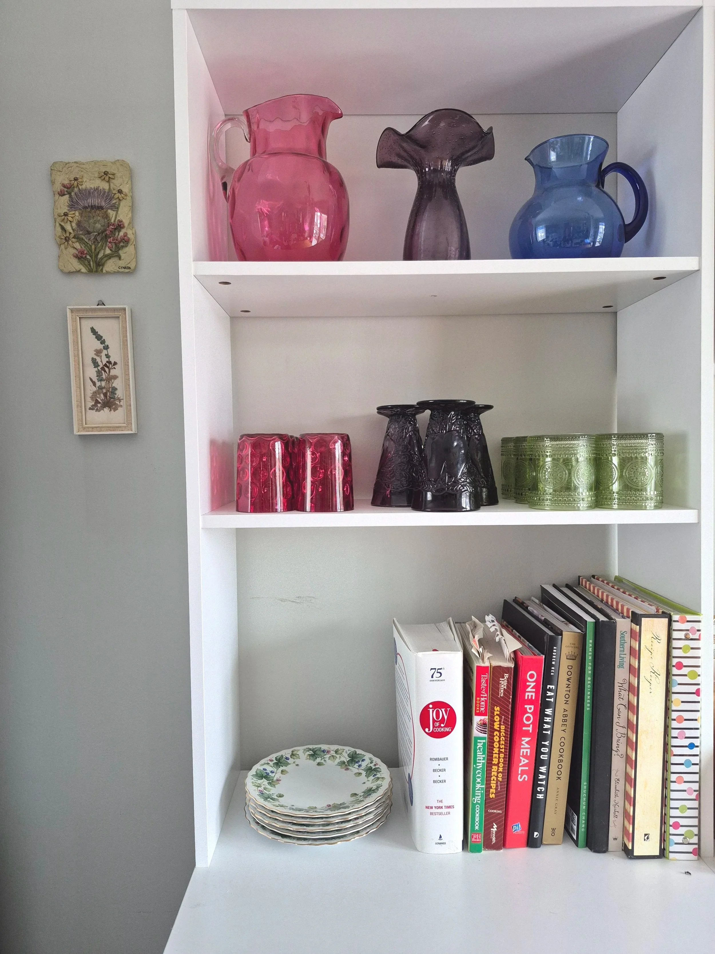 A bright white bookshelf has cranberry, purple, green, and cobalt glassware on the top two shelves, and a stack of floral plates and collection of cookbooks on the bottom shelf. Next to the bookshelf hang two small floral artworks.