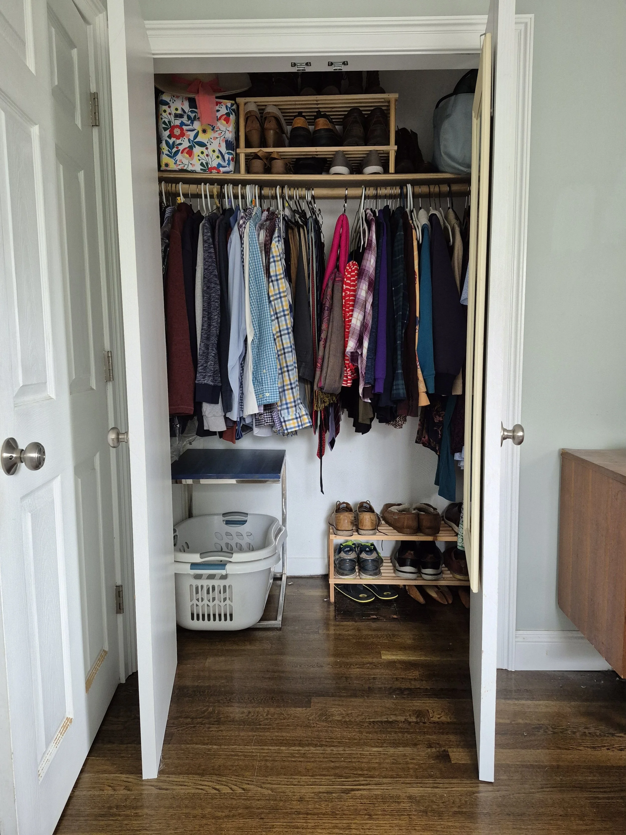 Clothes hanging in a closet. Below the clothes to the left is an empty laundry basket. To the right is a shoe rack. On the shelf above the hanging clothes is another shoe rack and storage bins and bags.