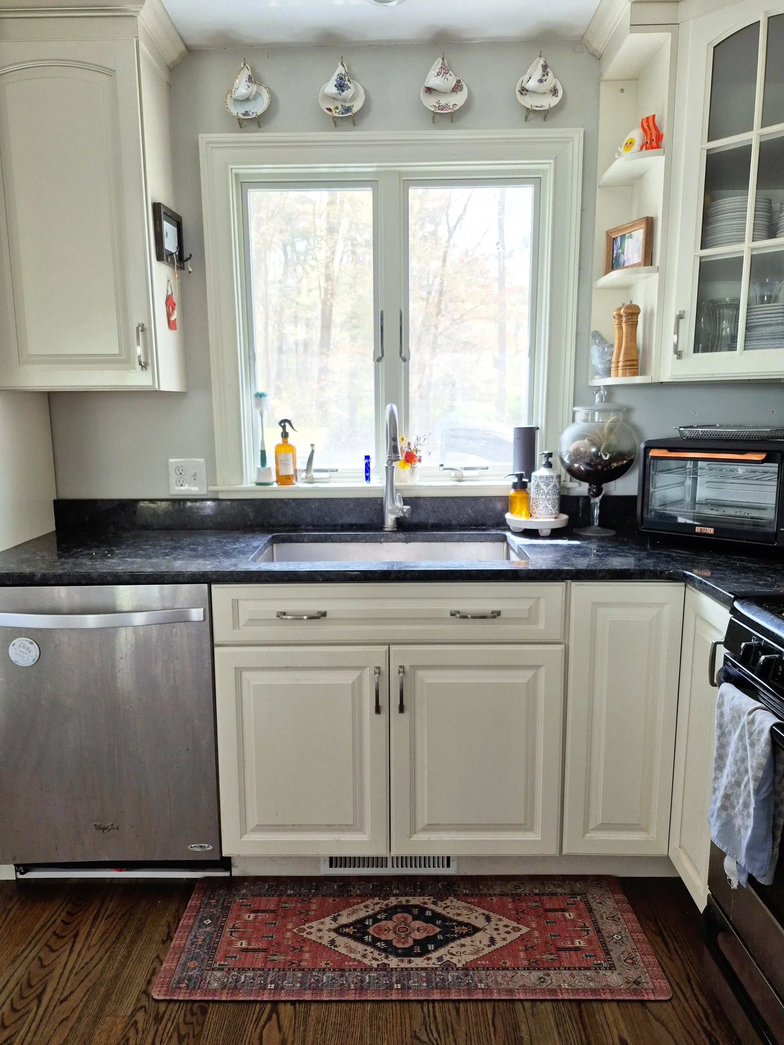 Teacups hang over a bright kitchen window. White cabinets and stainless steel appliances flank the kitchen sink under the window. Small knickknacks adorn the shelves next to the window.