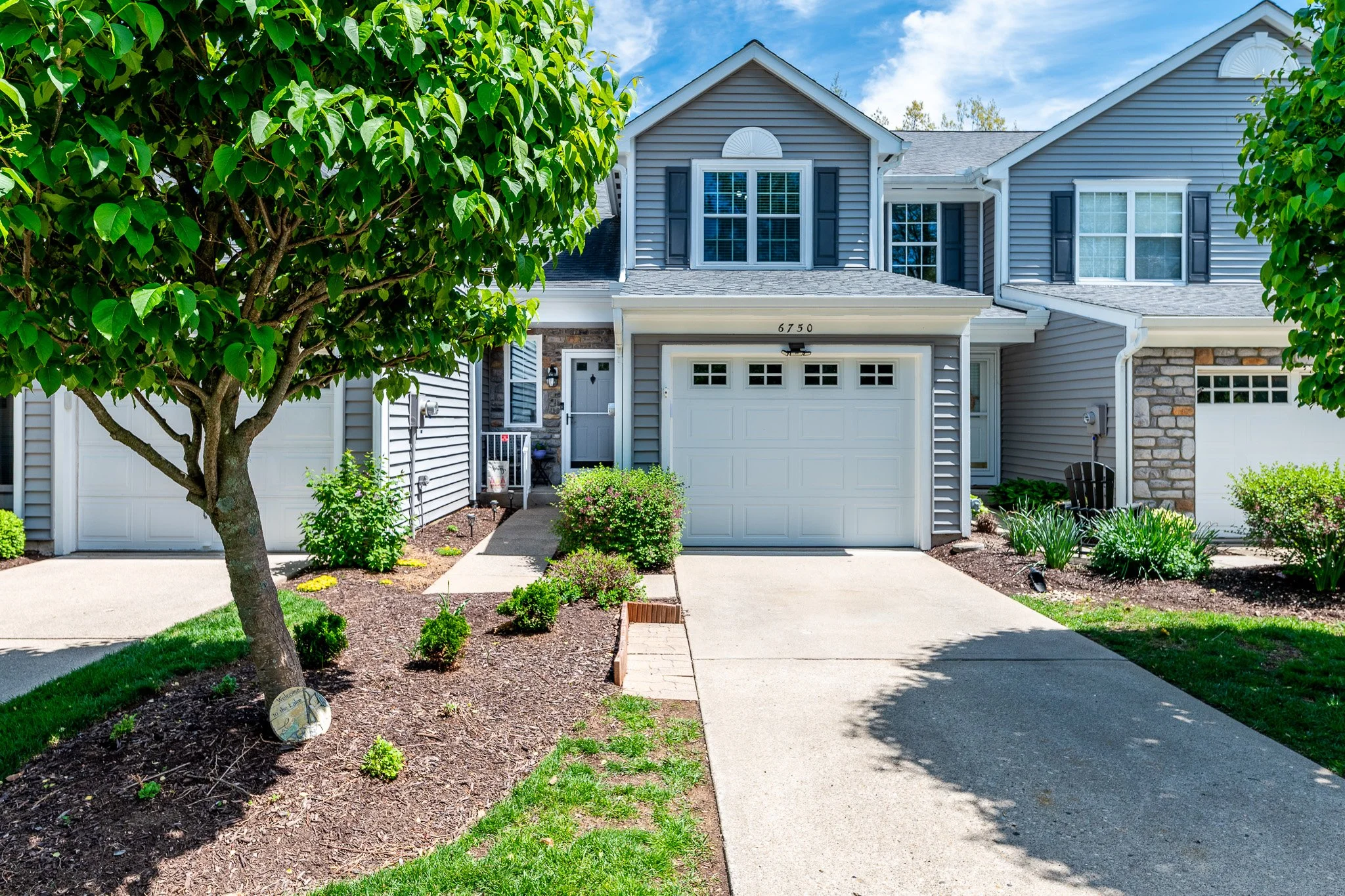 Front view of a two-story house with grey siding, white garage door, and black shutters on the windows. There is a driveway, a small pathway to the front door, and well-maintained landscaping with green bushes and trees.