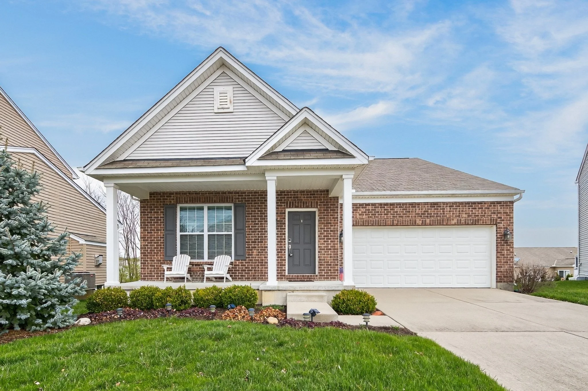 Front view of a suburban house with a brick exterior, white garage door, porch with two white chairs, and neatly landscaped front yard with bushes and grass.