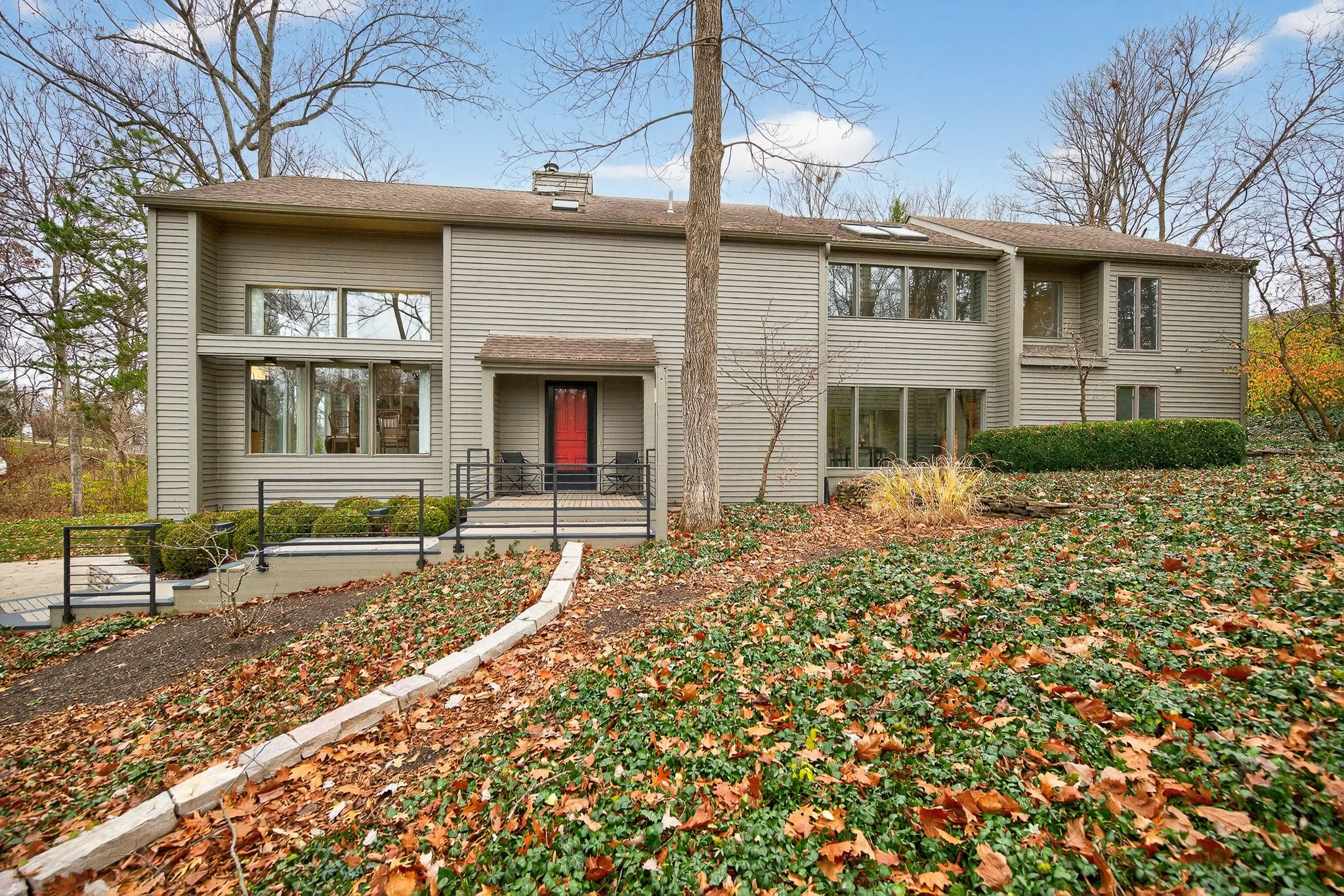 Multi-story house with beige siding, a red front door, and a small porch with black railing. The yard is filled with fallen leaves and some plants, with a large tree in front.