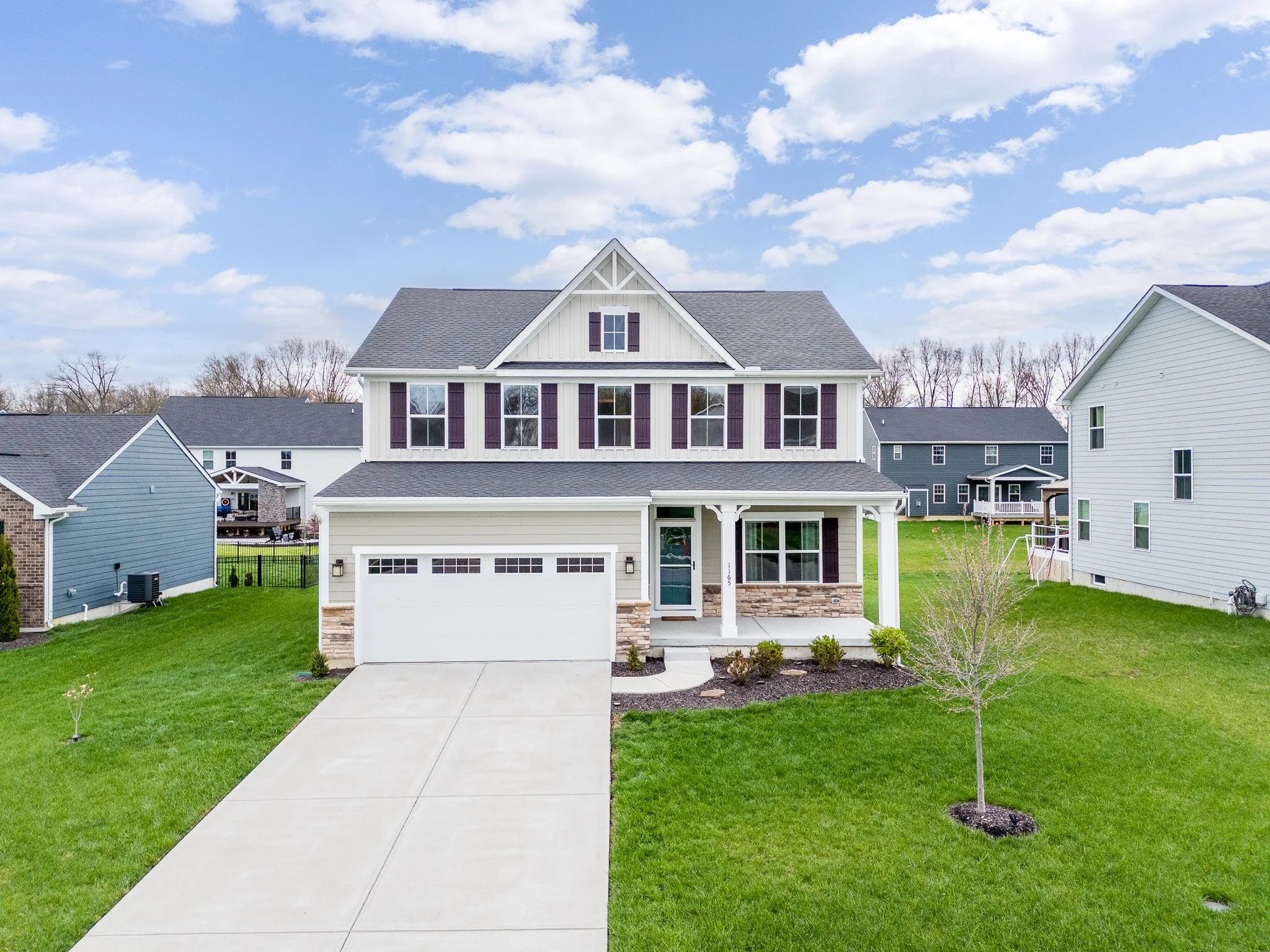 A two-story suburban house with a front porch, white siding, and black shutters, surrounded by green lawn and neighboring houses under a partly cloudy sky.