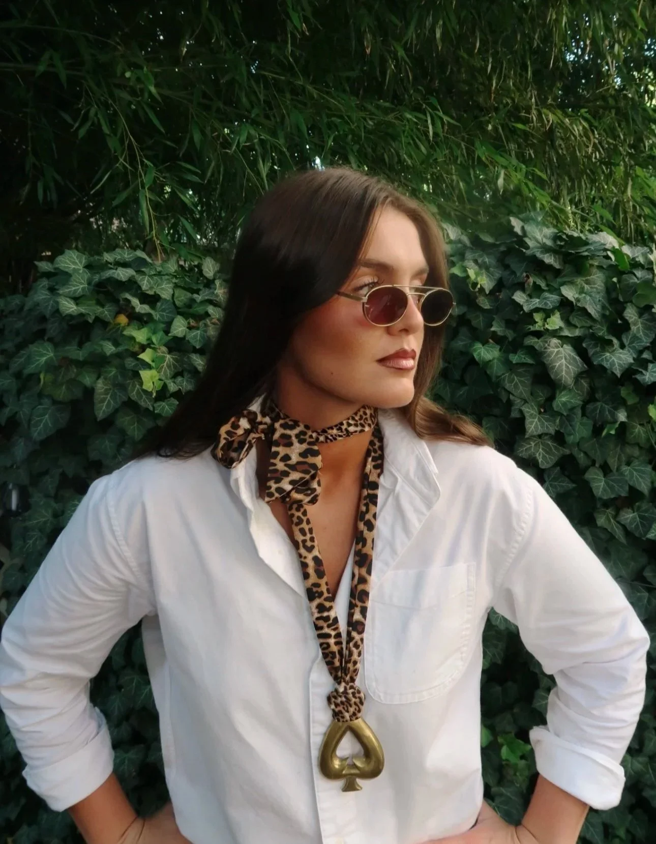 Woman wearing round sunglasses, leopard print scarf, white shirt, and a large gold spade pendant necklace, standing outdoors in front of green leafy bushes.