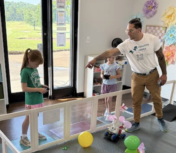 A man and two young girls in a room with glass door, with the man pointing at one girl who is wearing a green shirt and holding a device, and the other girl standing behind them smiling, with colorful paper flowers on the wall and balloons on the floor. Outside the door, a grassy area is visible.