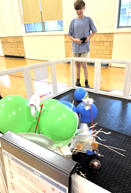 A boy laughing and watching a game involving an robot and balloons inside a gym or community center.
