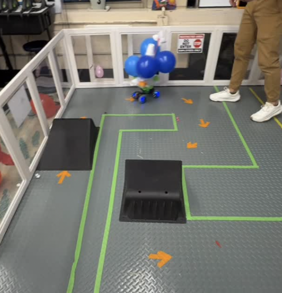 Robotic device with blue balloons on top, black ramps, orange arrows, and a person standing nearby in an enclosed area.