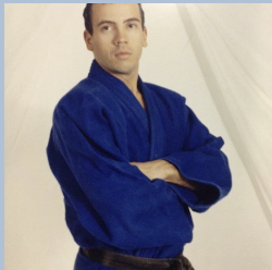 A man wearing a blue martial arts uniform with a black belt, standing with arms crossed against a light-colored background.