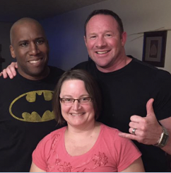 Three people smiling for a photo indoors. The person on the left is a man wearing a Batman t-shirt. The person in the middle is a woman with glasses wearing a pink top. The person on the right is a man with short hair wearing a black shirt and giving a thumbs-up.