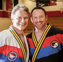 Two men wearing martial arts uniforms with medals, smiling indoors.