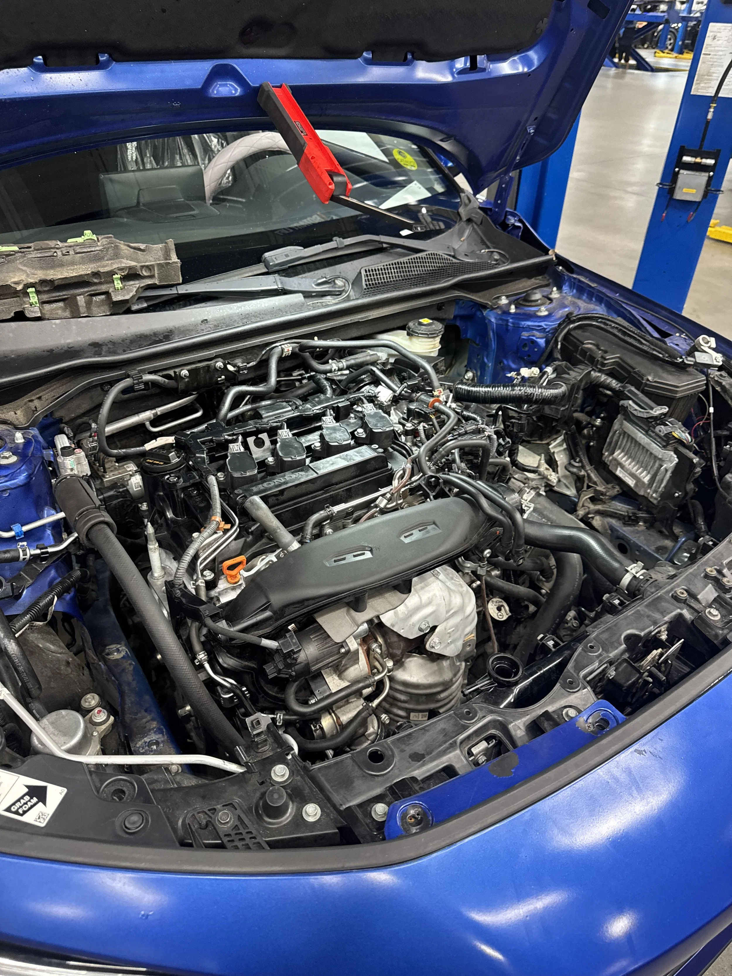 The image shows the open hood of a blue car, exposing the engine and various mechanical components inside a garage or workshop setting.