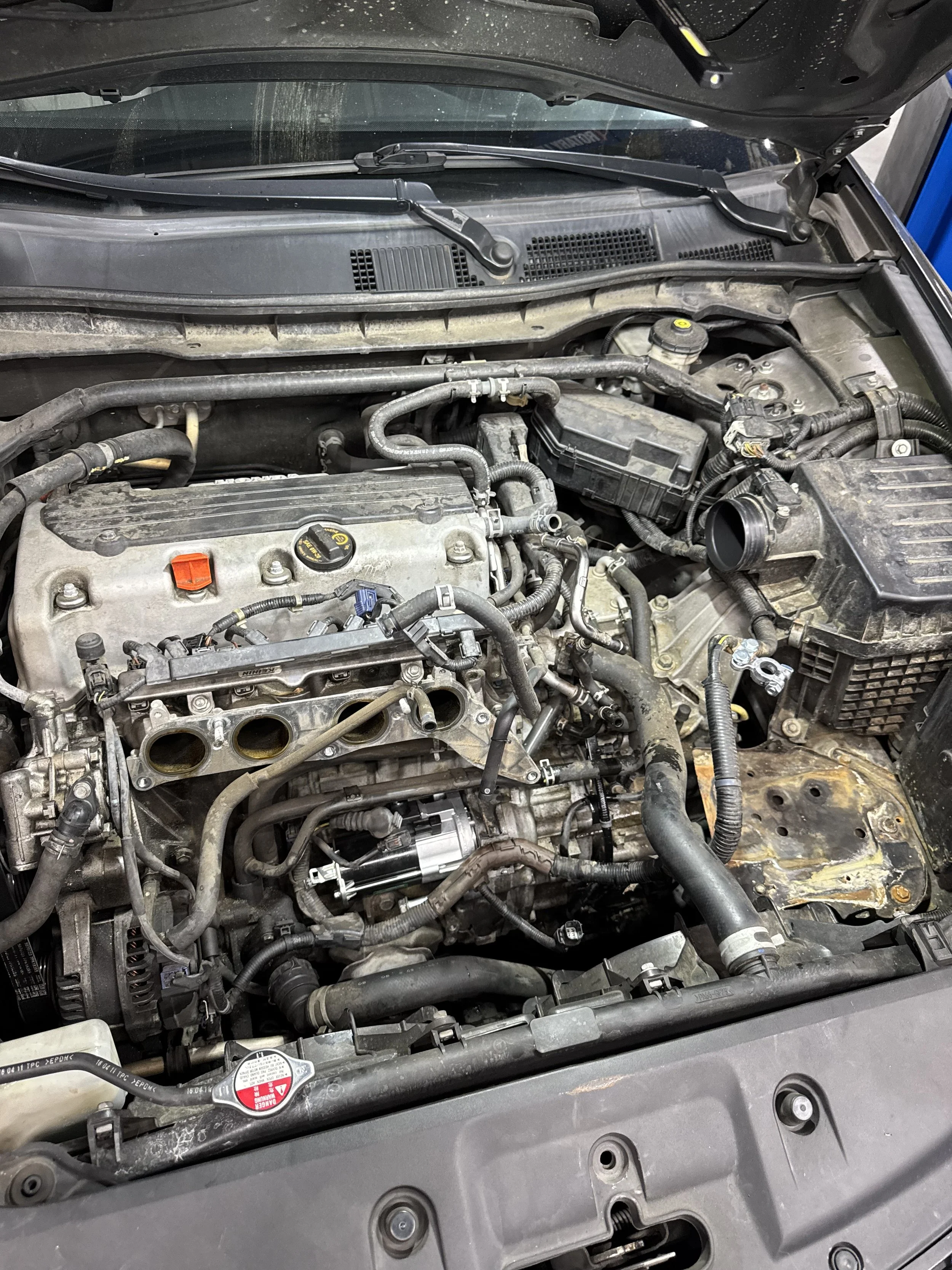 View of a car's open hood showing engine components, hoses, and parts covered in dust and dirt.