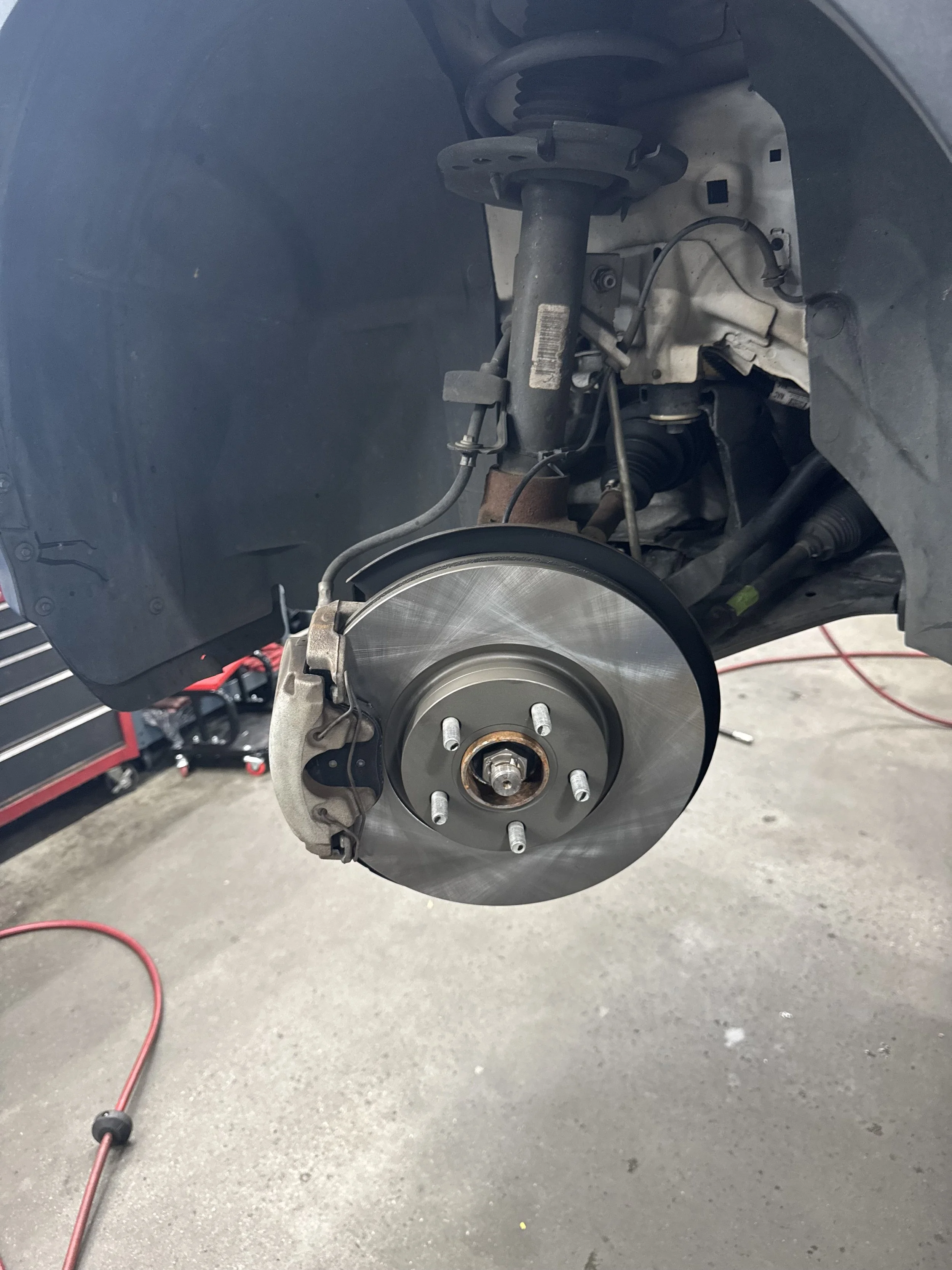Close-up of a car's front wheel area with the wheel removed, showing the brake rotor, brake caliper, and suspension components in a repair shop.