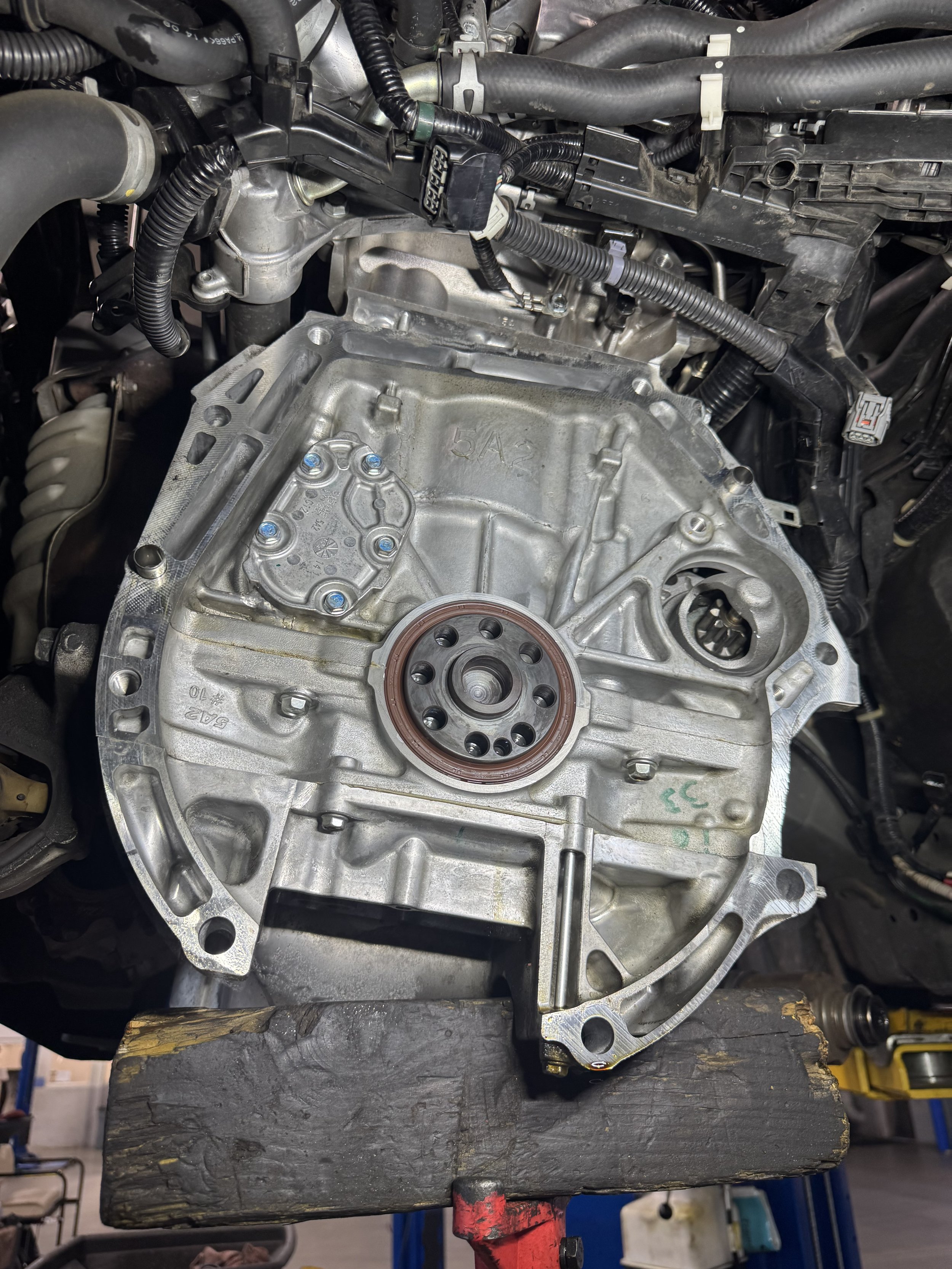 Automobile engine transmission housing with visible mounting points and internal components, placed on a wooden block in a workshop.