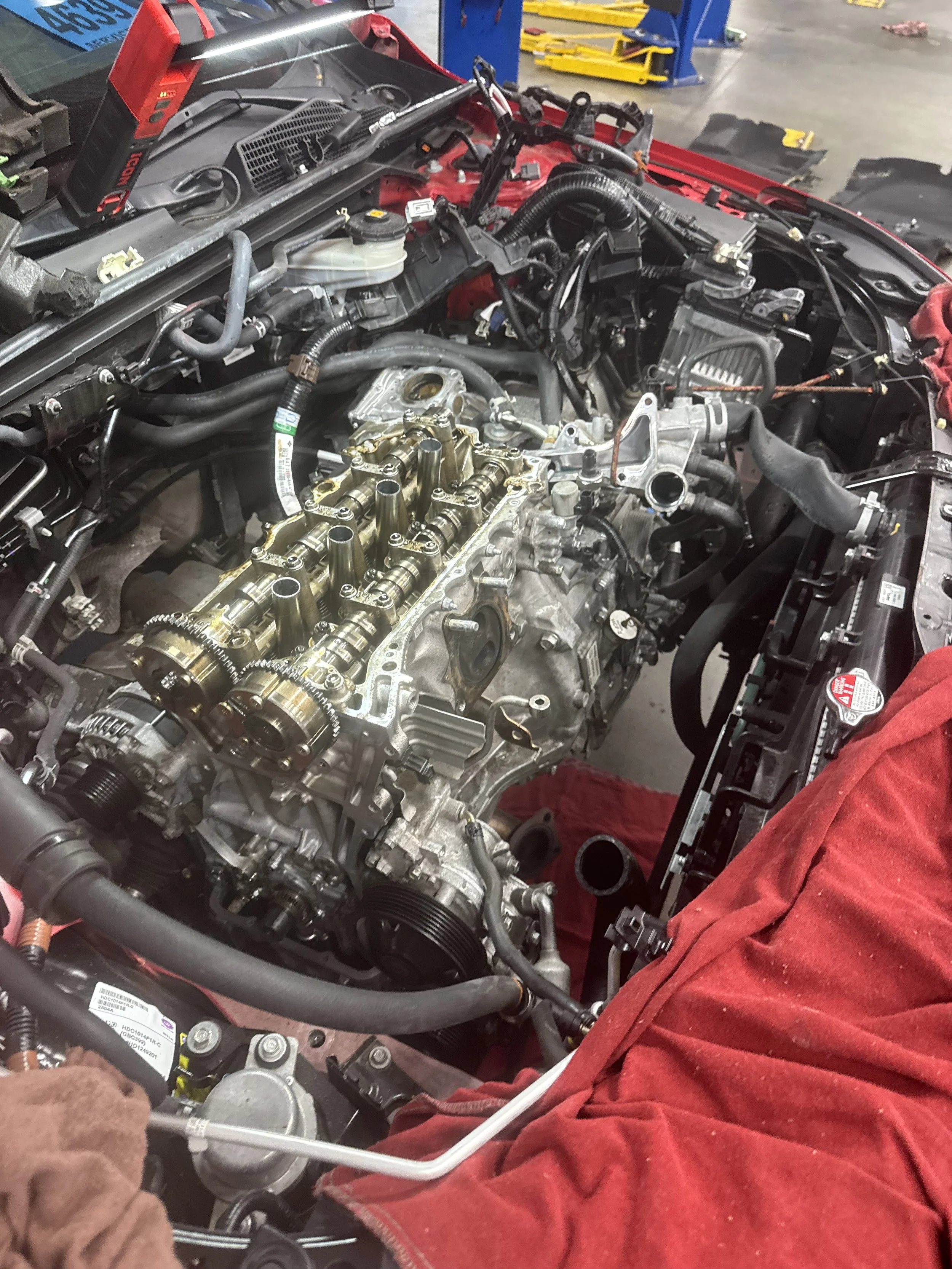 A car engine with the cylinder head removed, exposing the camshafts and valves, in a garage or workshop setting.