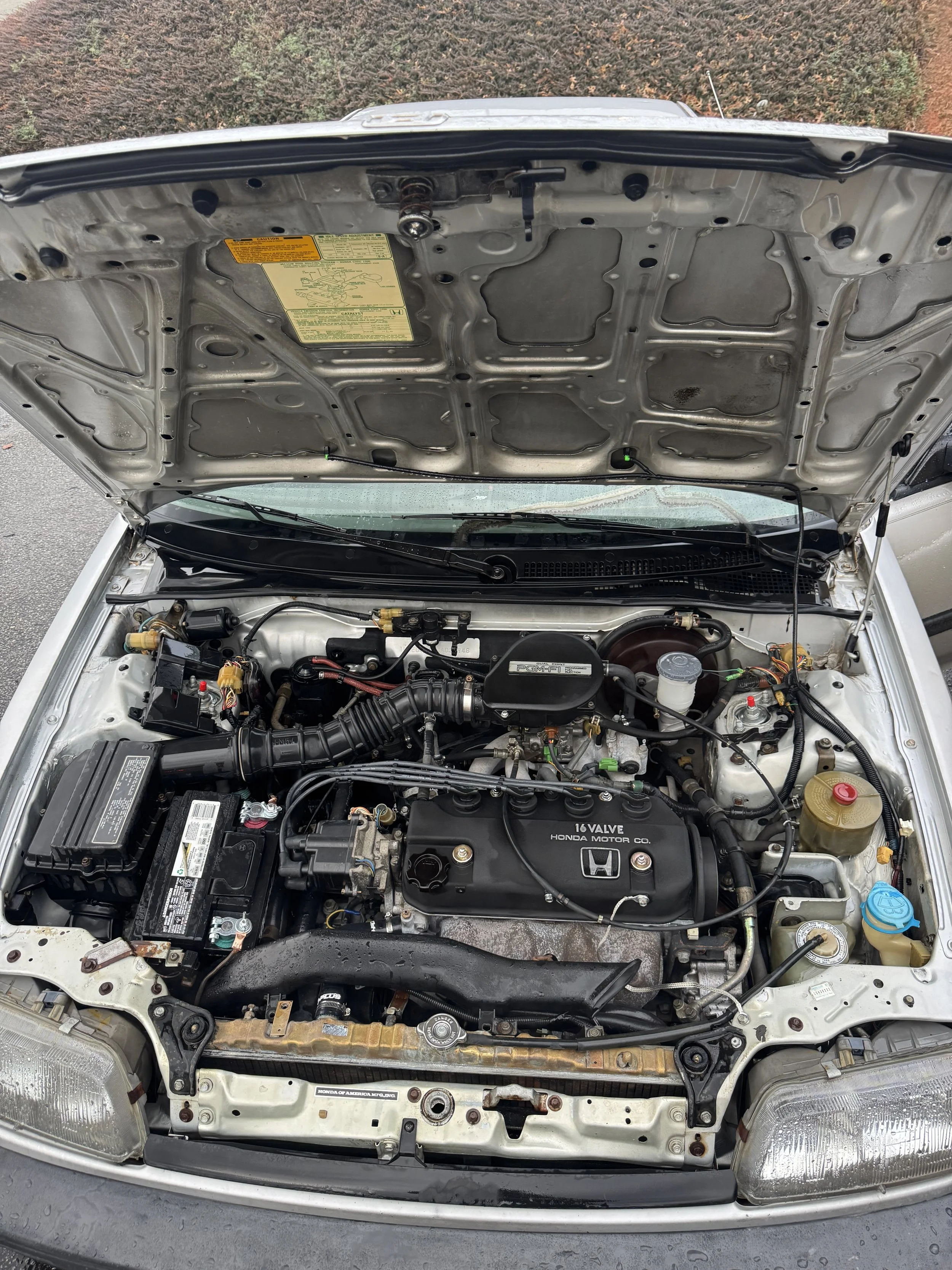 Open hood of a car showing the engine bay with a Honda engine, car battery, radiator, and various engine components.