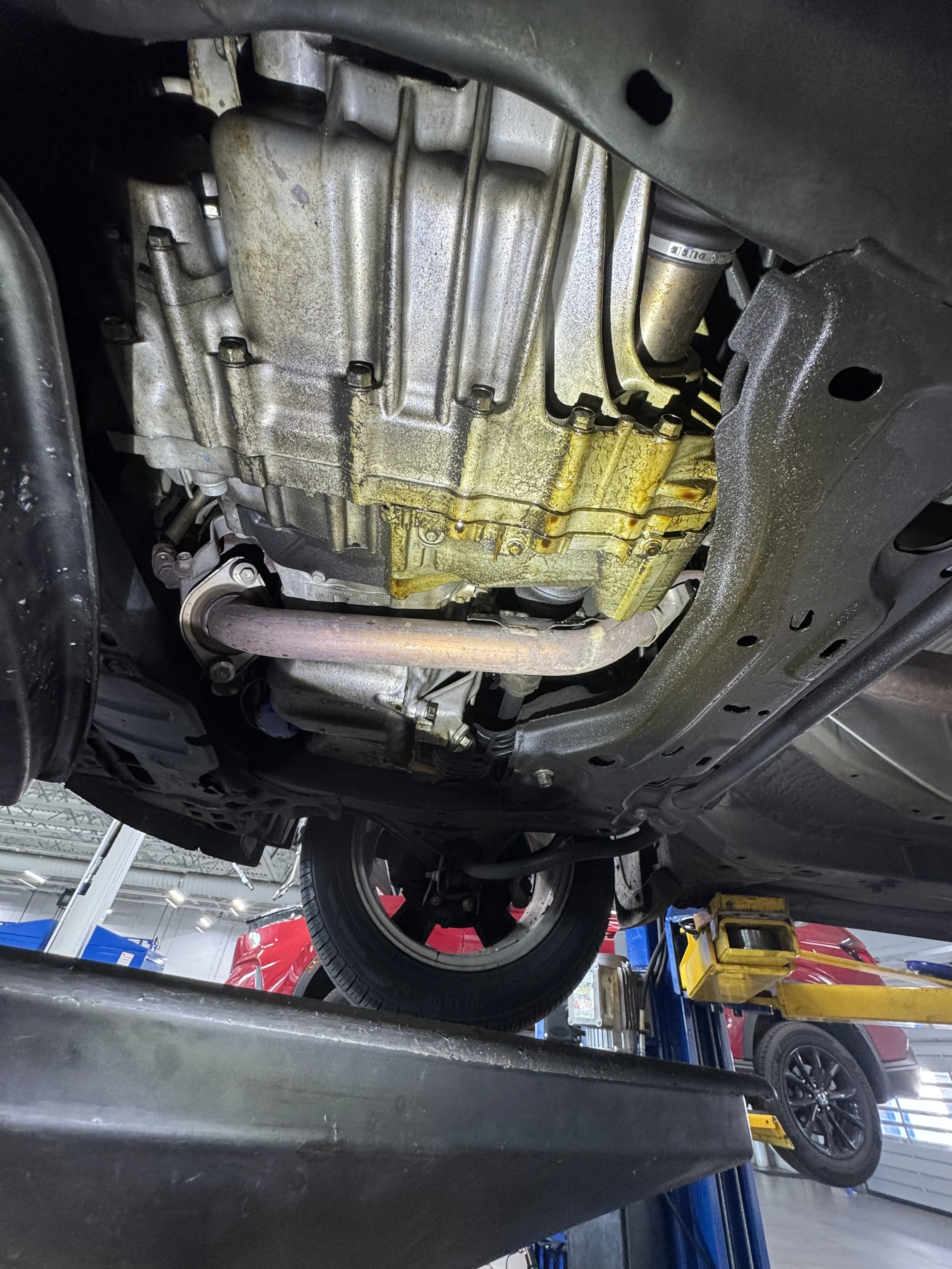 Underneath view of a car's engine with oil leakage, visible oil stains, and a part of the exhaust pipe, lifted on a hydraulic lift in an automotive repair shop.