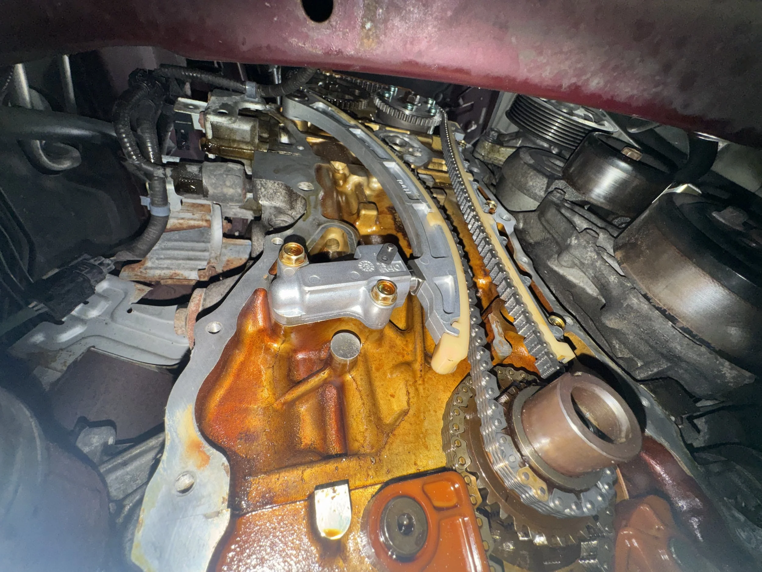 Close-up of a car engine with the valve cover removed, exposing the timing chain, sprockets, and internal components, which are partially covered in engine oil and rust.