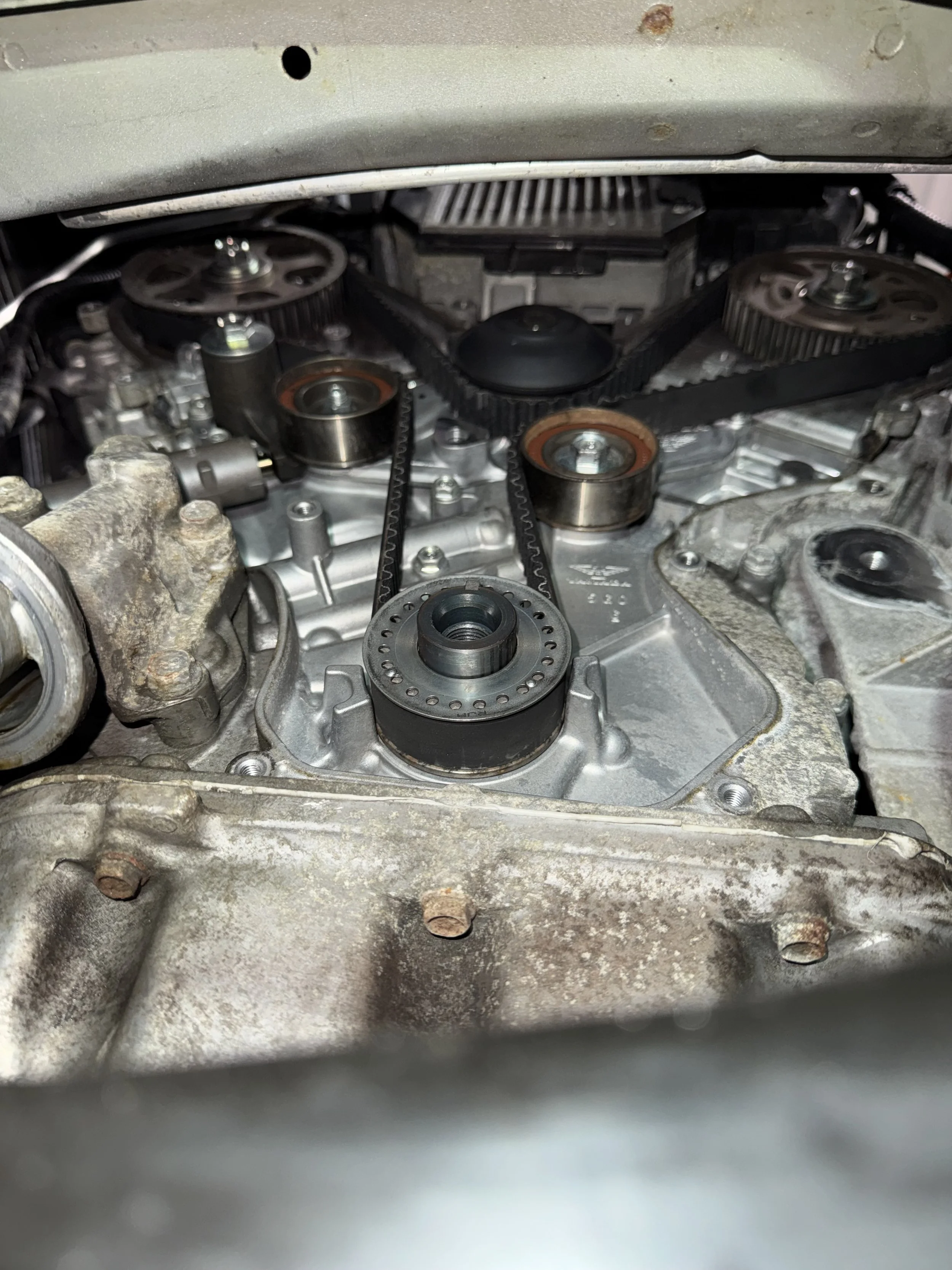 Close-up of a car engine with a timing belt, pulleys, and metal parts, showing some rust and dirt.