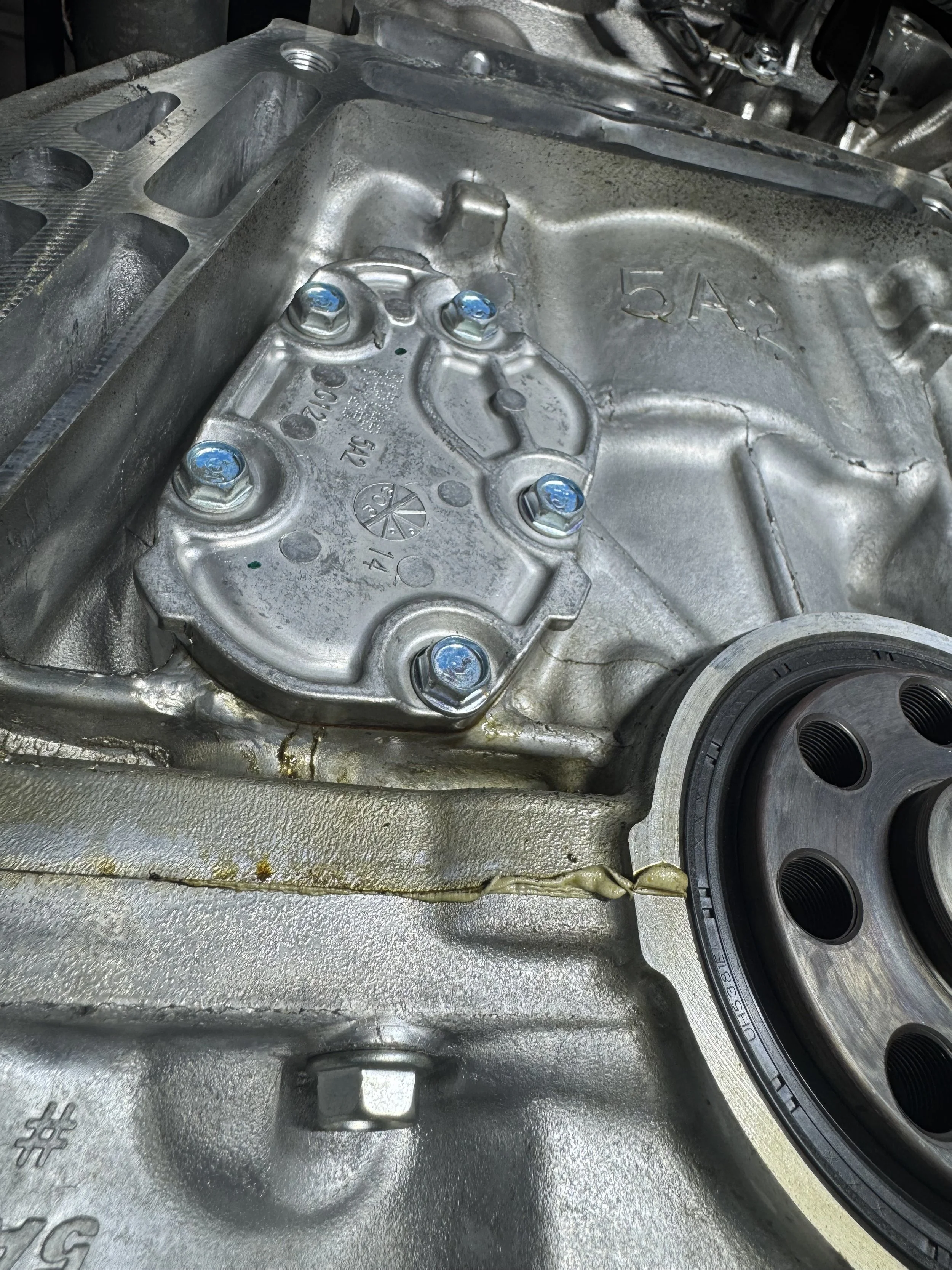 View of an engine block with a silver metal cover, several bolts, and part of a black pulley with holes.