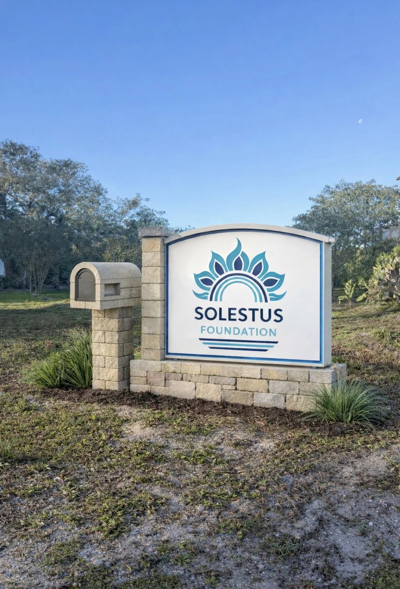 Sign for Solestus Foundation with a stylized lotus or sunburst logo, surrounded by small bushes and a mailbox, under a clear blue sky with some trees in the background.