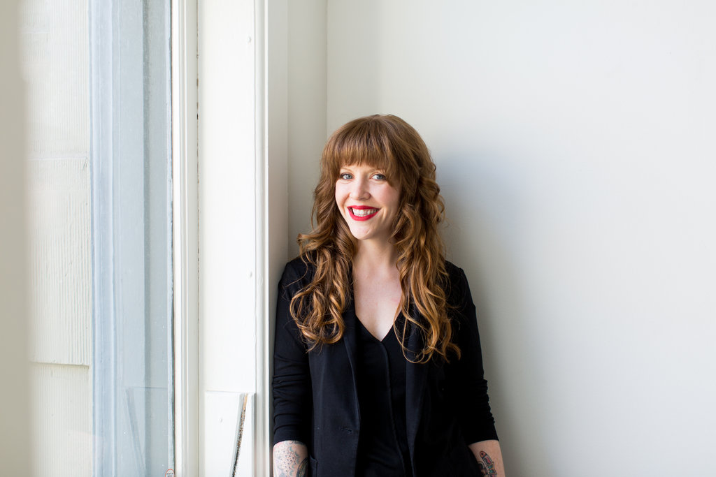 A woman with long, curly red hair standing near a window, smiling, wearing a black blazer and top.
