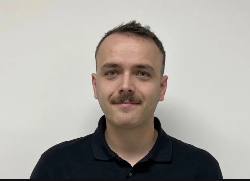 A young man with short brown hair, a mustache, and light skin, wearing a black polo shirt, standing against a white wall and smiling.