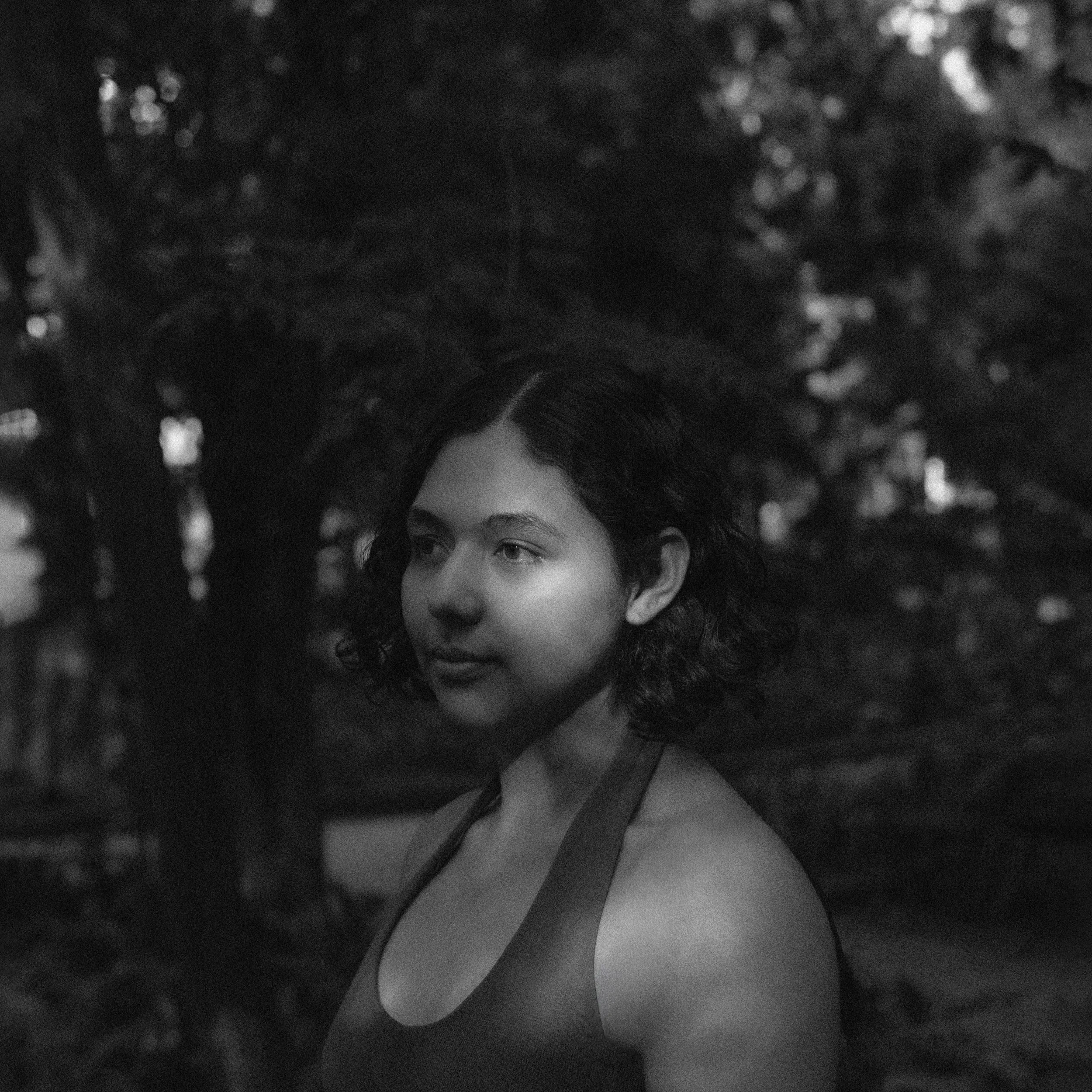 Black and white photo of a young woman with short curly hair, standing outdoors with trees in the background.