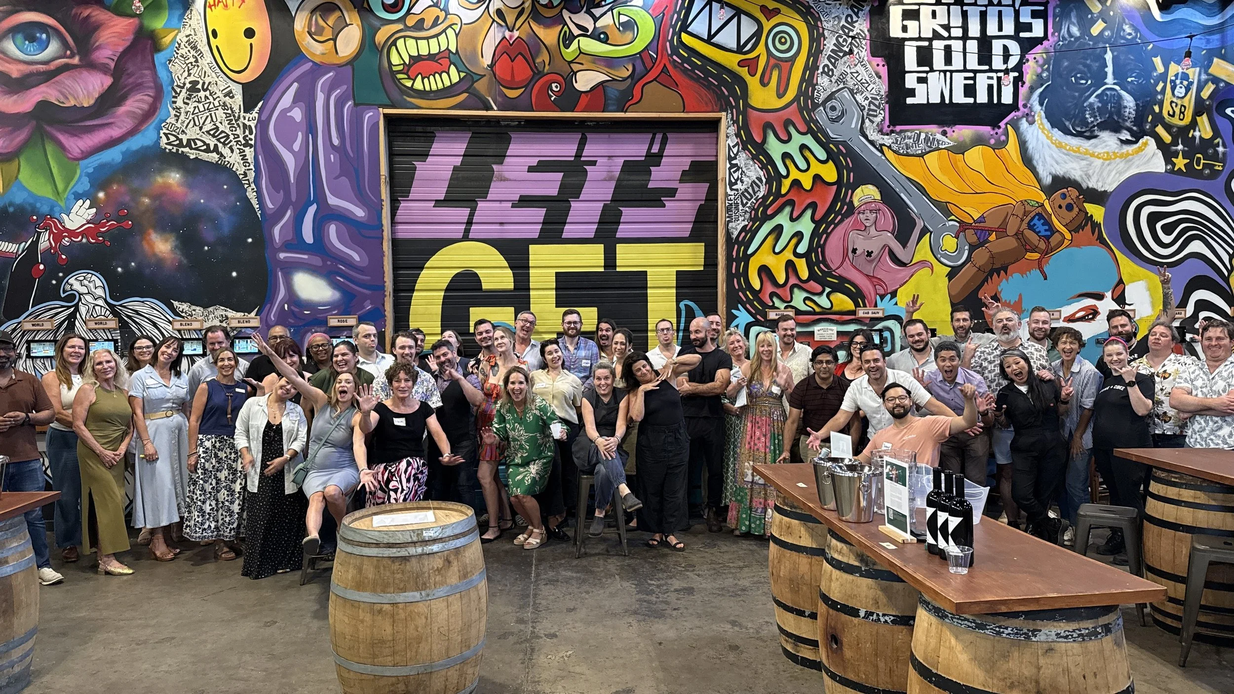 A large group of people gathered indoors, smiling and posing for a photo in front of colorful graffiti art on the walls, with some people raising their hands and making playful gestures.