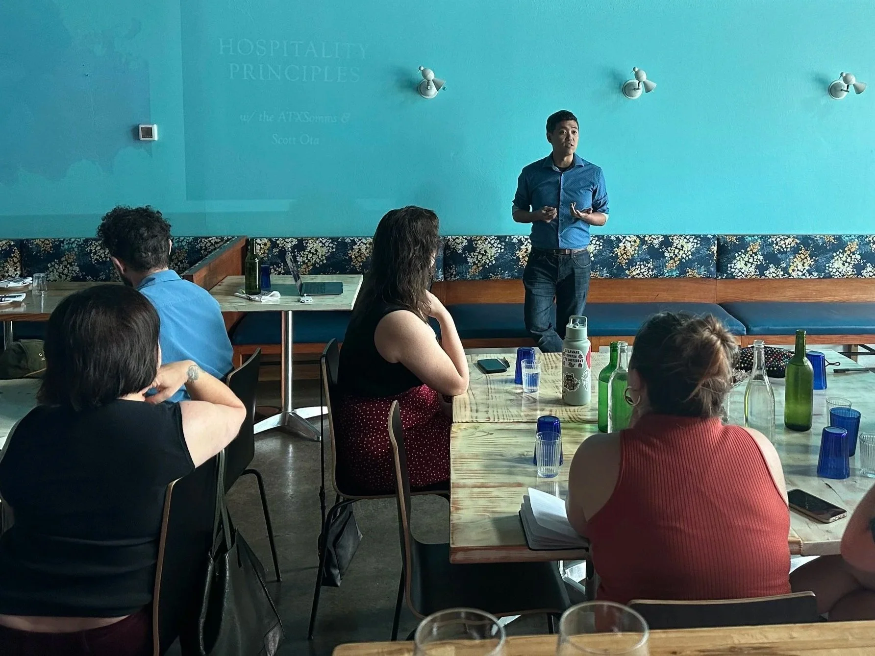A man in a blue shirt giving a presentation to a small group of people seated at a table with drinks and notebooks in a restaurant or cafe with teal walls and floral patterned seating.