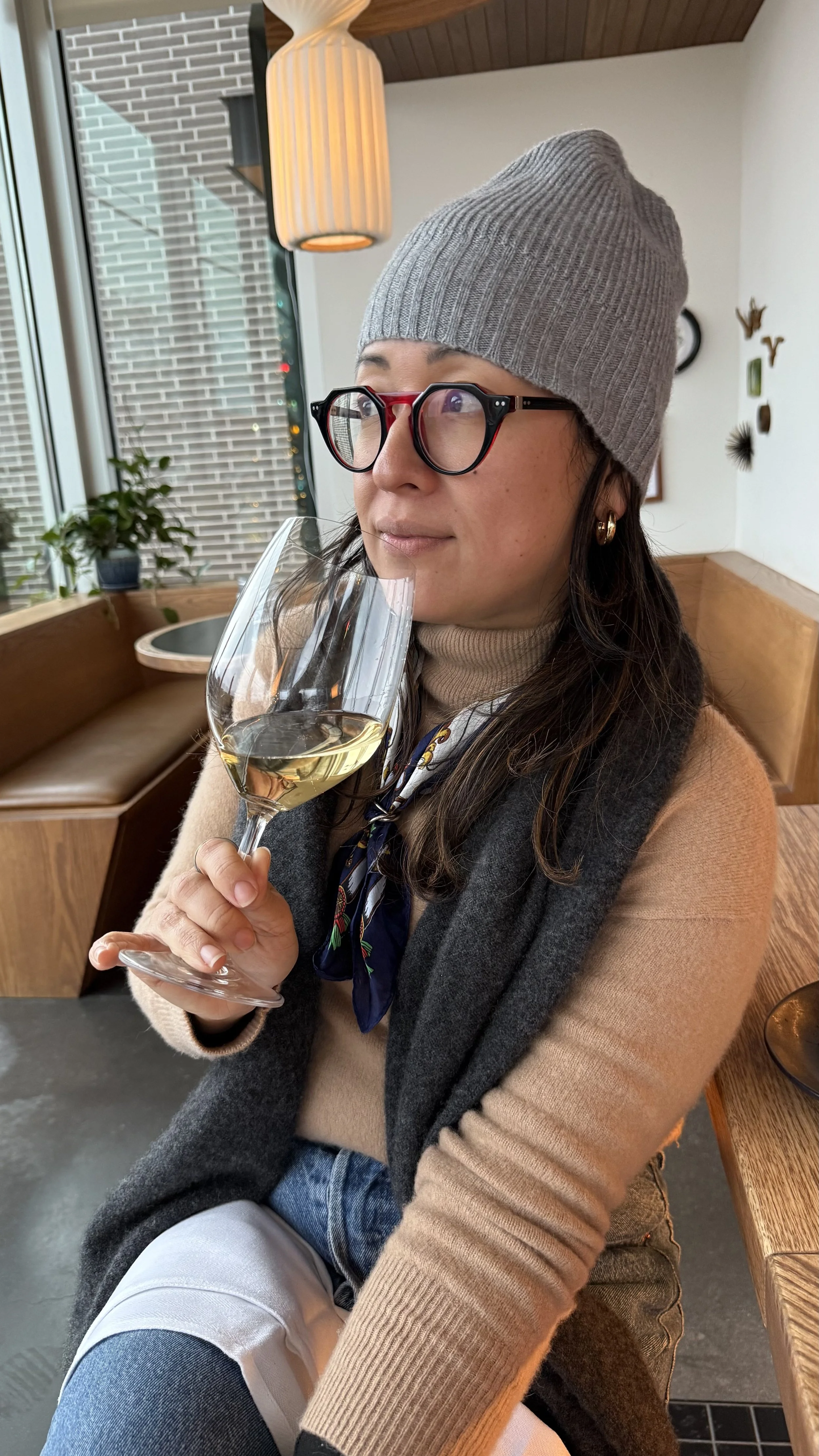 A woman wearing glasses, a gray beanie, a beige turtleneck, and a gray vest, sitting indoors near a window with a wine glass of white wine, with modern decor and plants in the background.