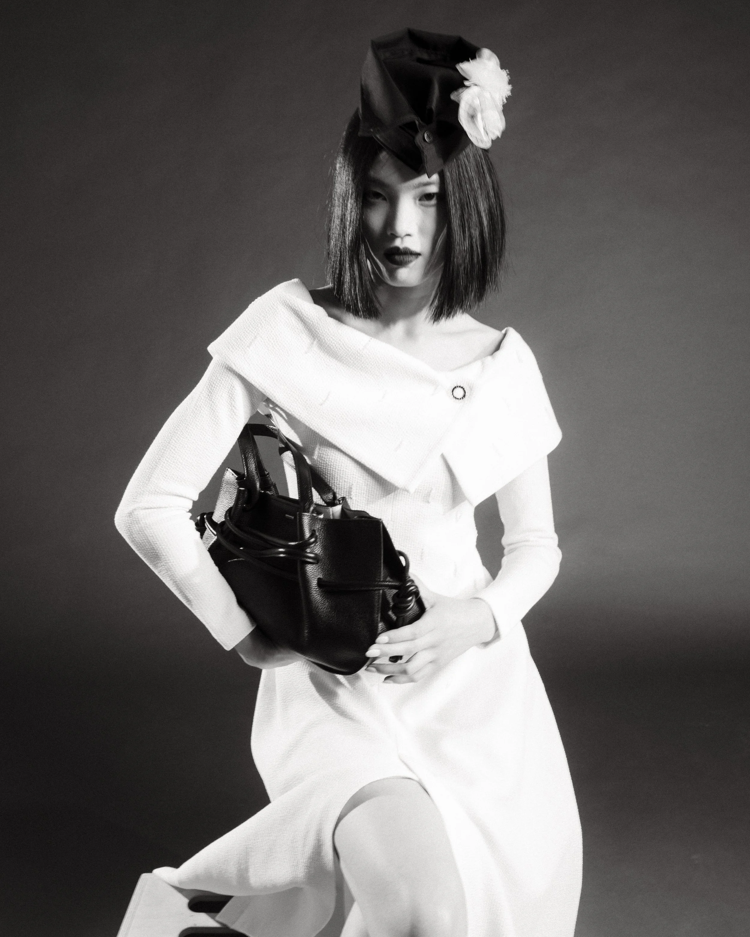 Black and white photo of a woman with shoulder-length dark hair, wearing a white off-the-shoulder dress, a black hat with a flower adornment, holding a black handbag and looking at the camera.