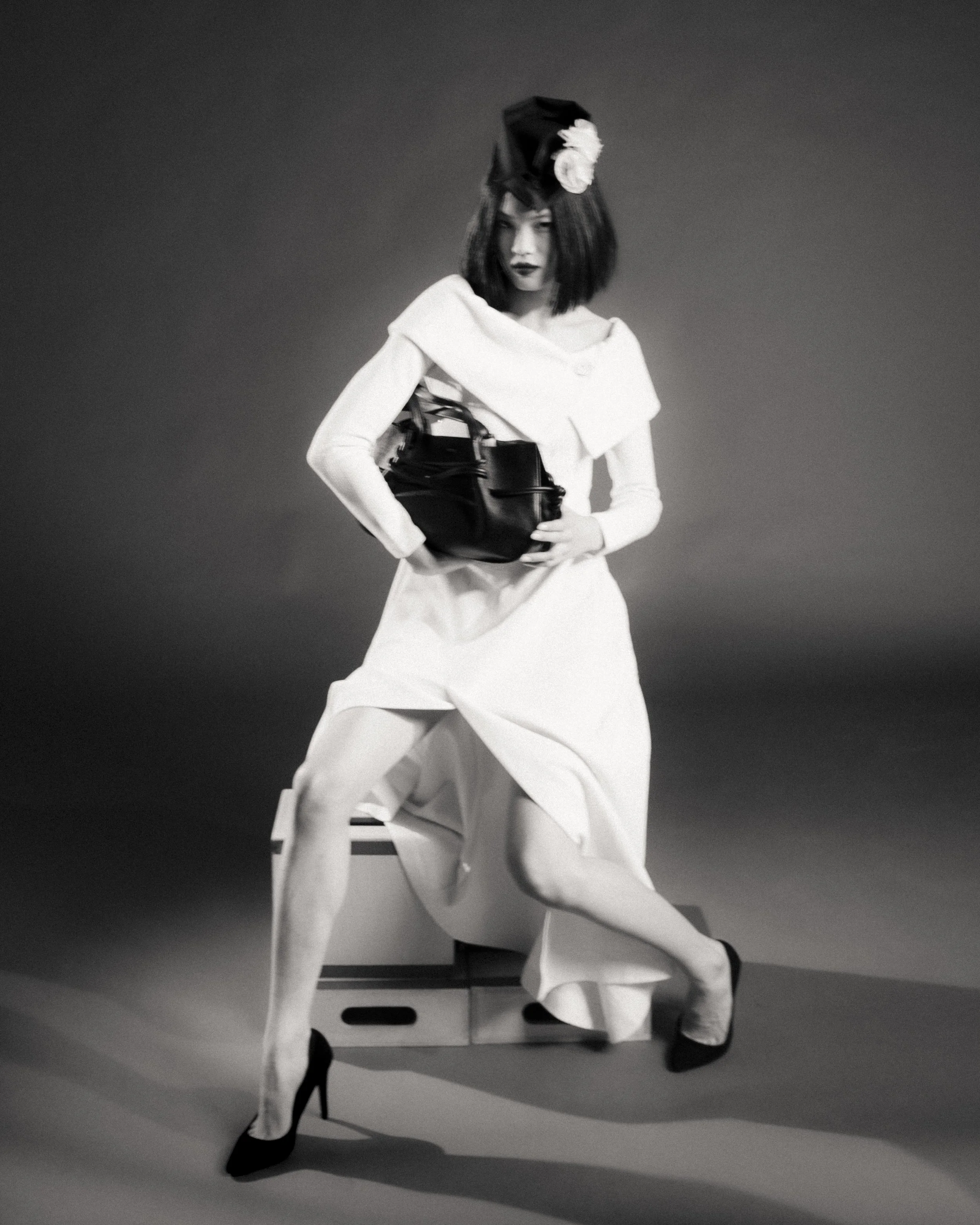 Black and white photograph of a woman with dark lipstick and short hair, wearing a large hat with a flower, a white blouse, and wide-legged pants, holding a black bag, standing against a plain background.
